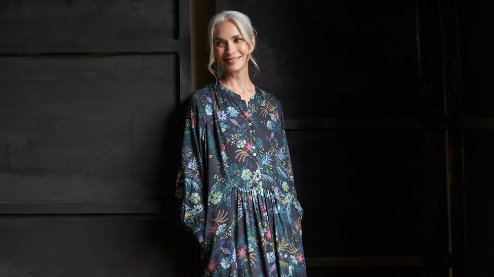 An older woman in a floral dress leaning against a wall.
