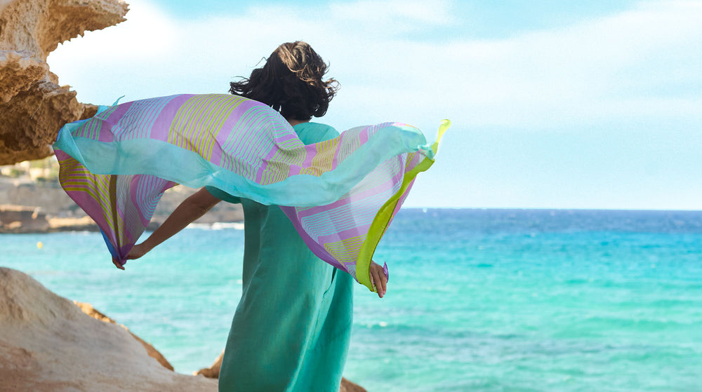 A person standing by the sea holding a colorful scarf fluttering in the breeze.