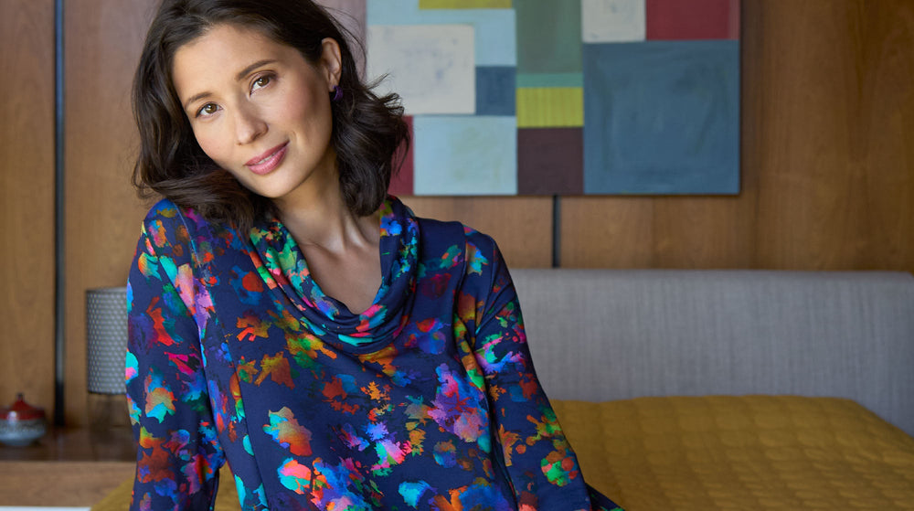 Woman in a colorful shirt and white pants lying on a beige surface against a blue background, looking upwards with one arm behind her head.