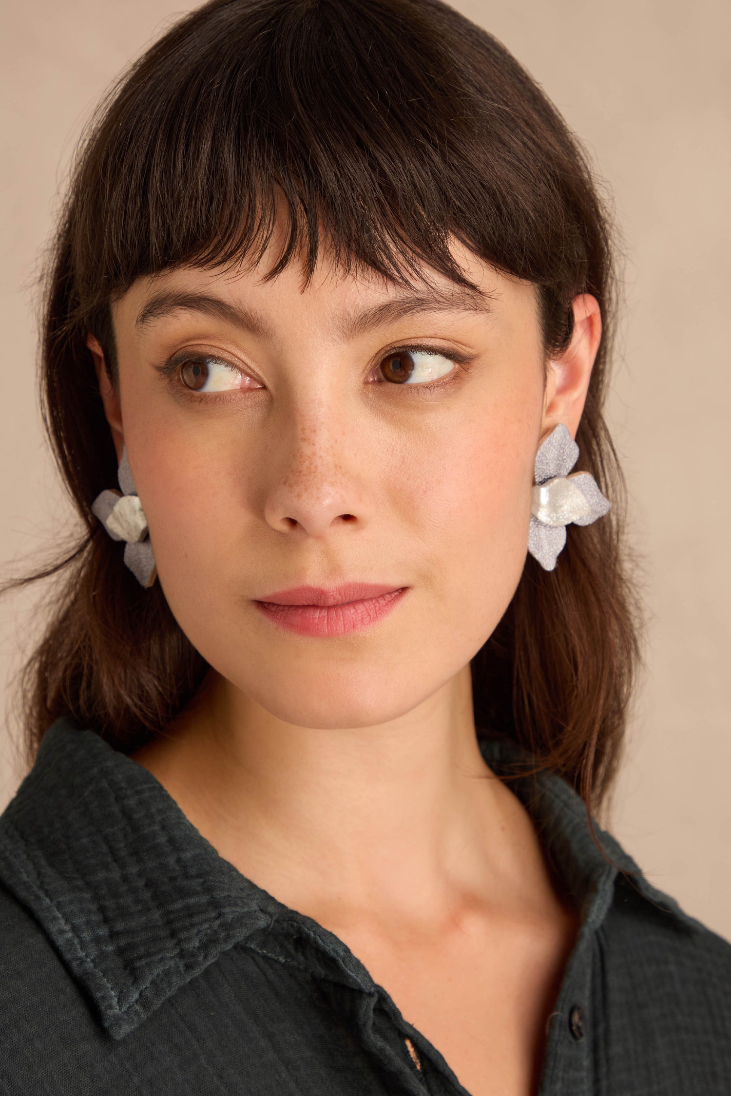 Woman with straight brown hair and bangs wears Metallic Grey Leather Petal Earrings and a dark collared shirt, looking slightly left against a neutral background.
