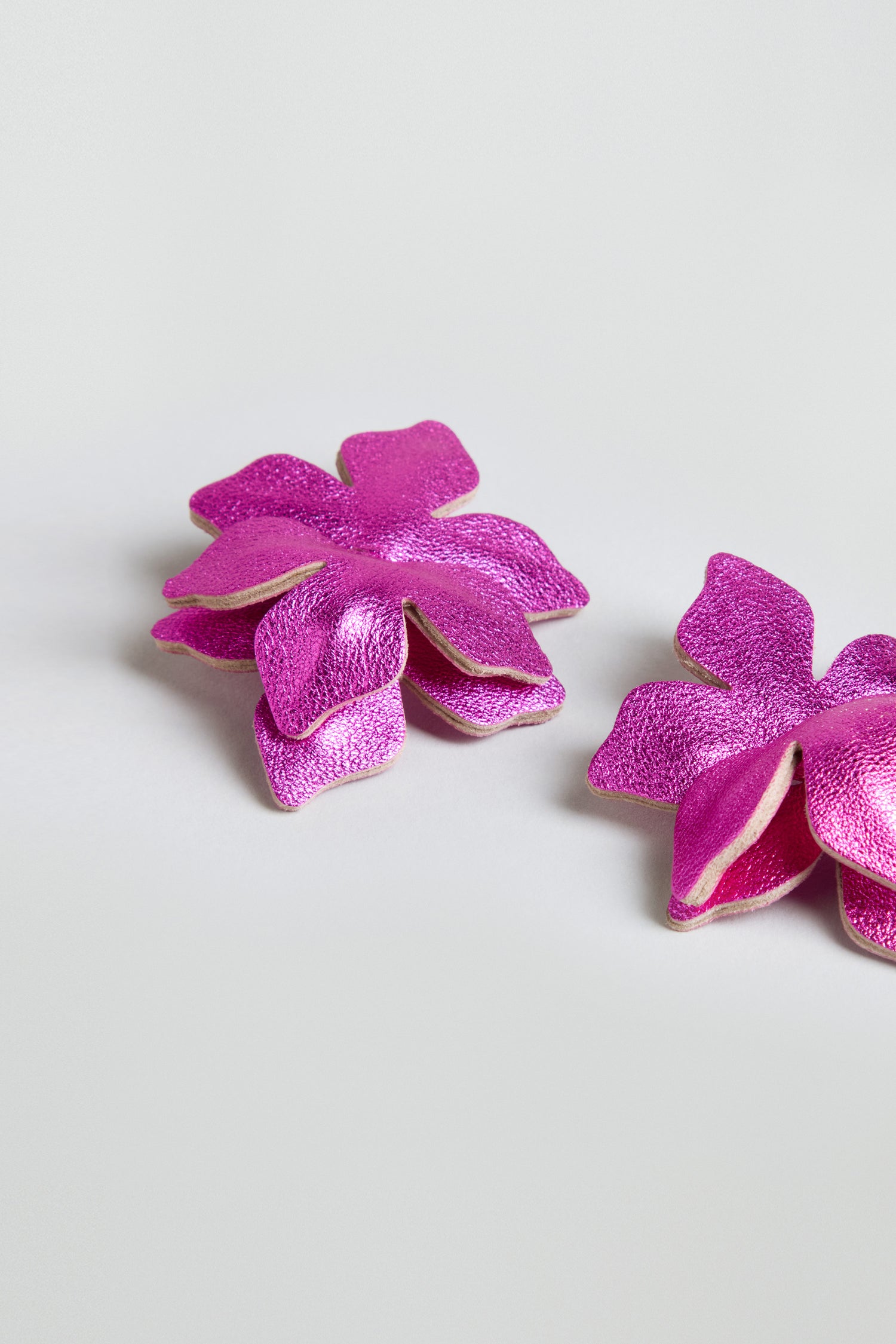 A pair of Metallic Leather Flower Earrings in fuchsia, featuring layered floral shapes, are displayed on a simple light gray surface.