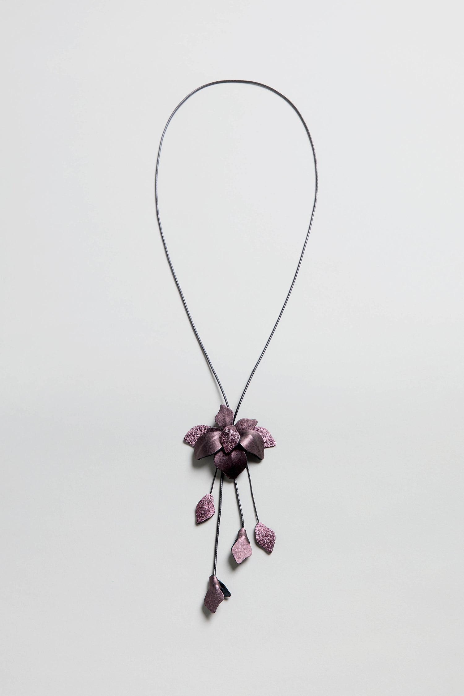 The Metallic Leather Flower Necklace features a purple flower pendant and five petal-like charms on a thin metallic leather chain, shown on a white background.