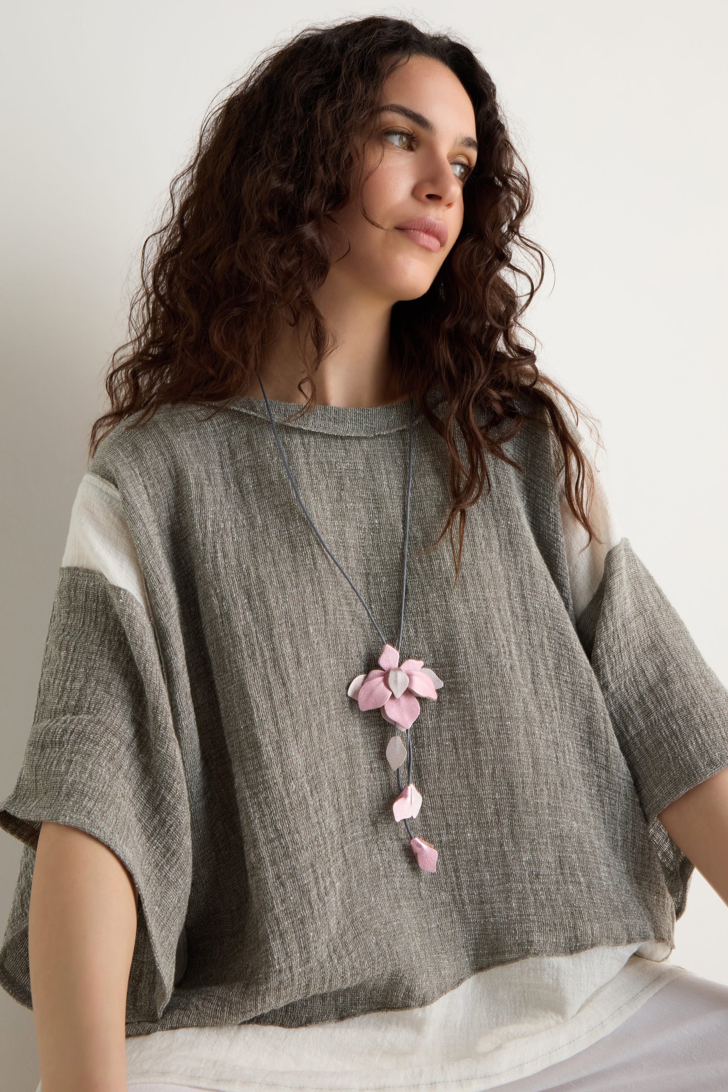 Woman with wavy brown hair wears a loose grey and white top and a long cord necklace featuring the Leather Flower Pendant, which has a handcrafted pink flower, while seated against a plain light background.