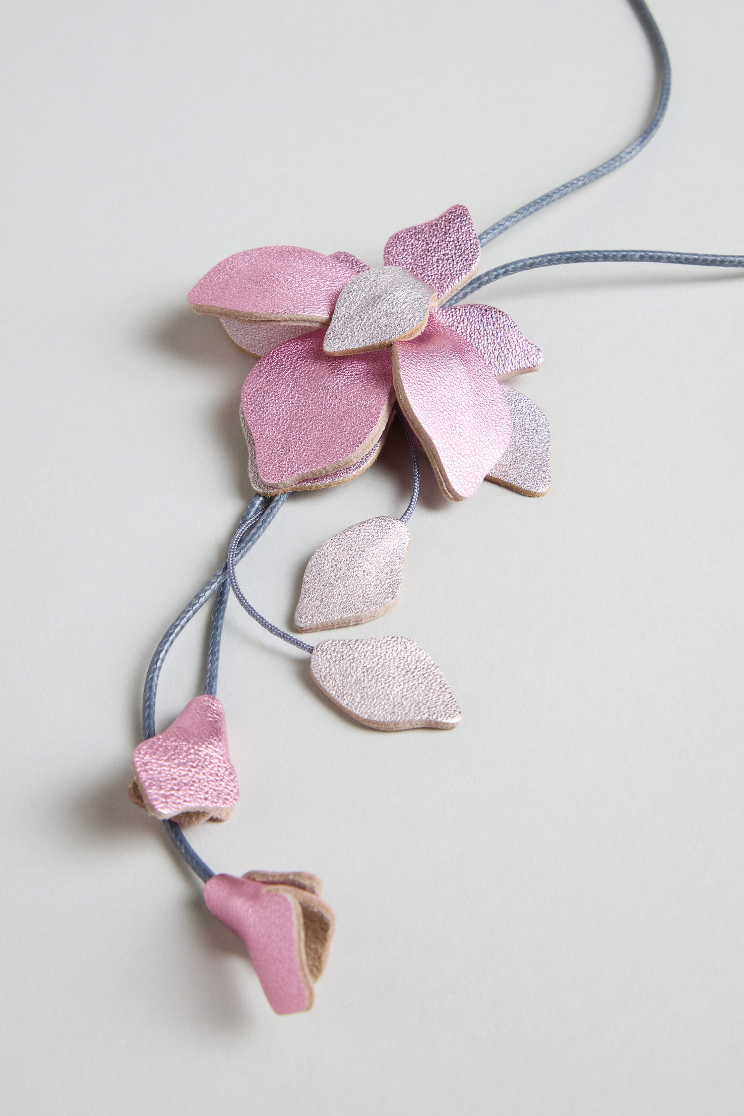 The Leather Flower Pendant is a handcrafted necklace with pink and lilac leather shaped like a blossom with leaves, featuring a long cord and shown against a light grey background.