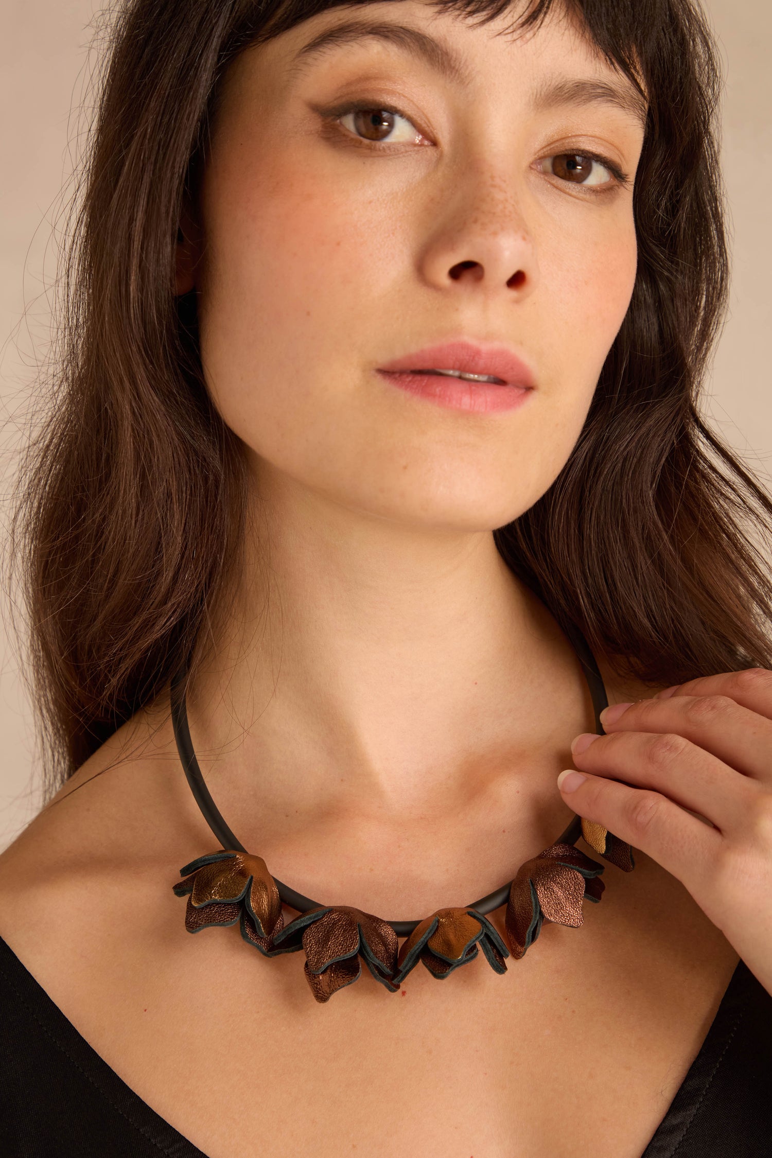 Woman with long dark hair in a black top wears the Short Metallic Leather Petals Necklace, featuring brown leaf-shaped pendants, and looks at the camera neutrally.