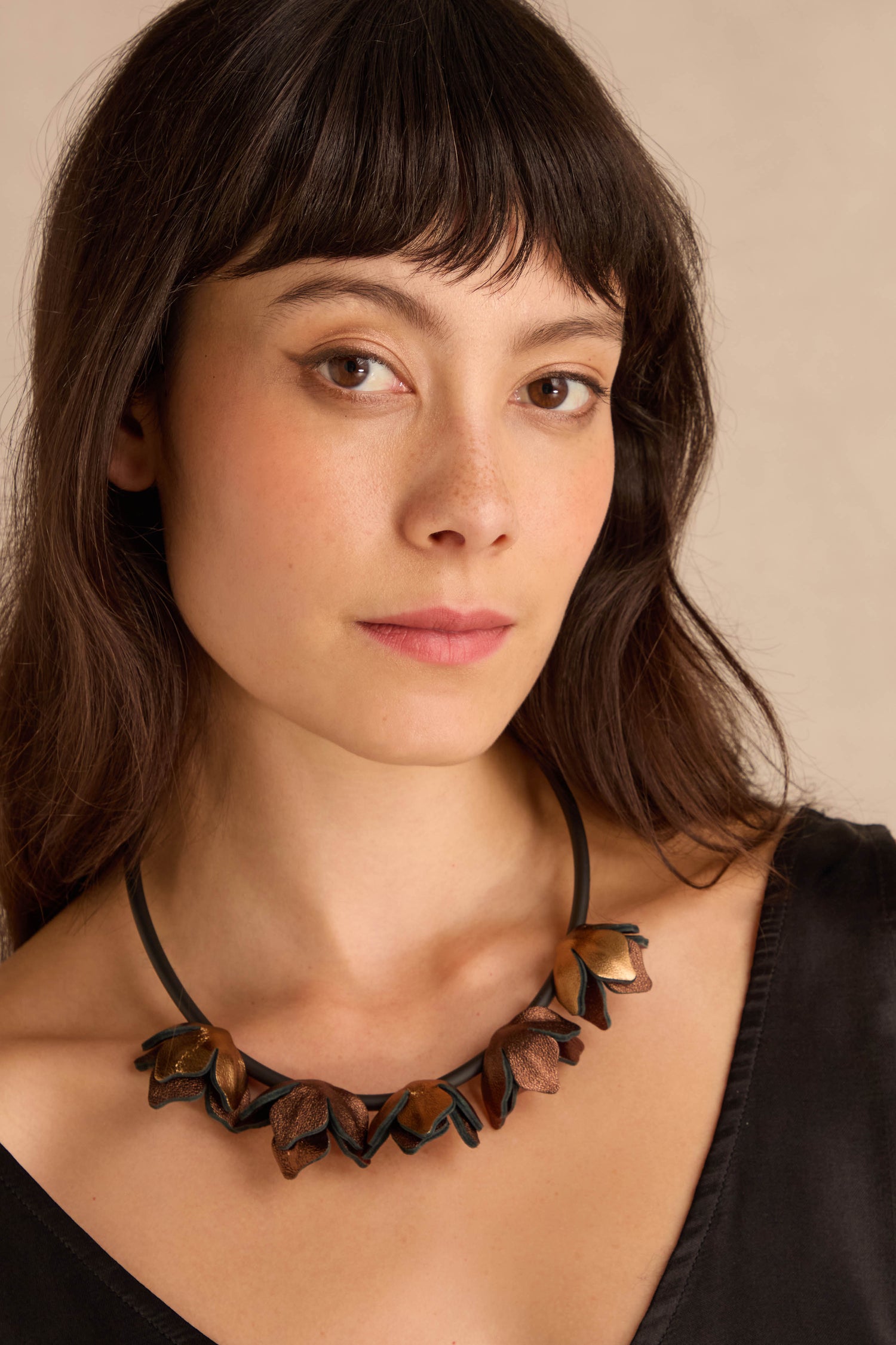 Woman with long brown hair wears a black top and the Short Metallic Leather Petals Necklace in burgundy and bronze, posing against a plain beige background.