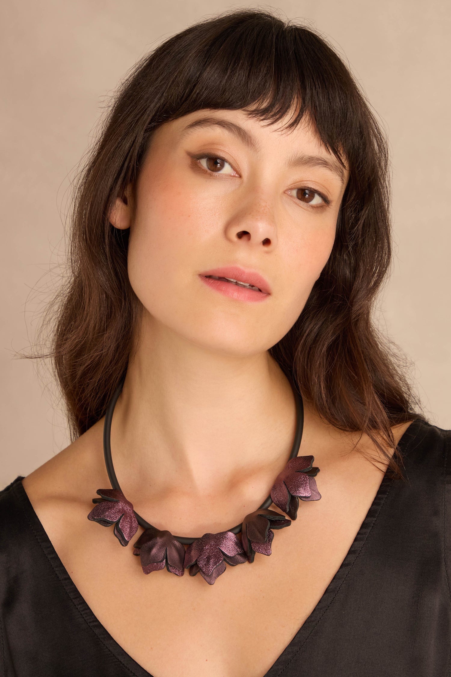 Woman with straight dark hair wears a black V-neck top and the Short Metallic Leather Petals Necklace with purple flower shapes, looking slightly upward against a beige background.