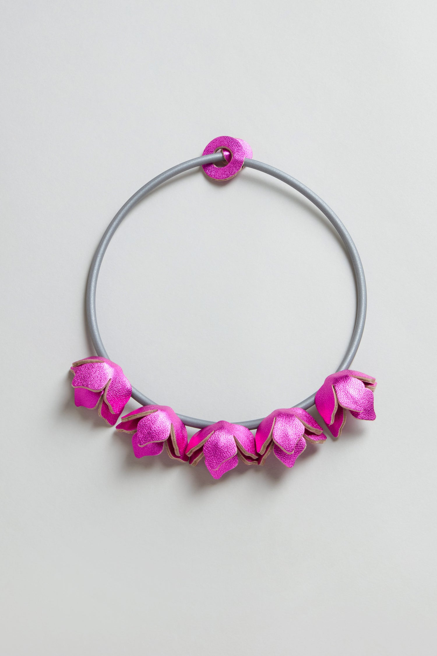 The Short Leather Petals Necklace features a circular gray band with five evenly spaced metallic fuchsia flower-shaped decorations along the bottom.