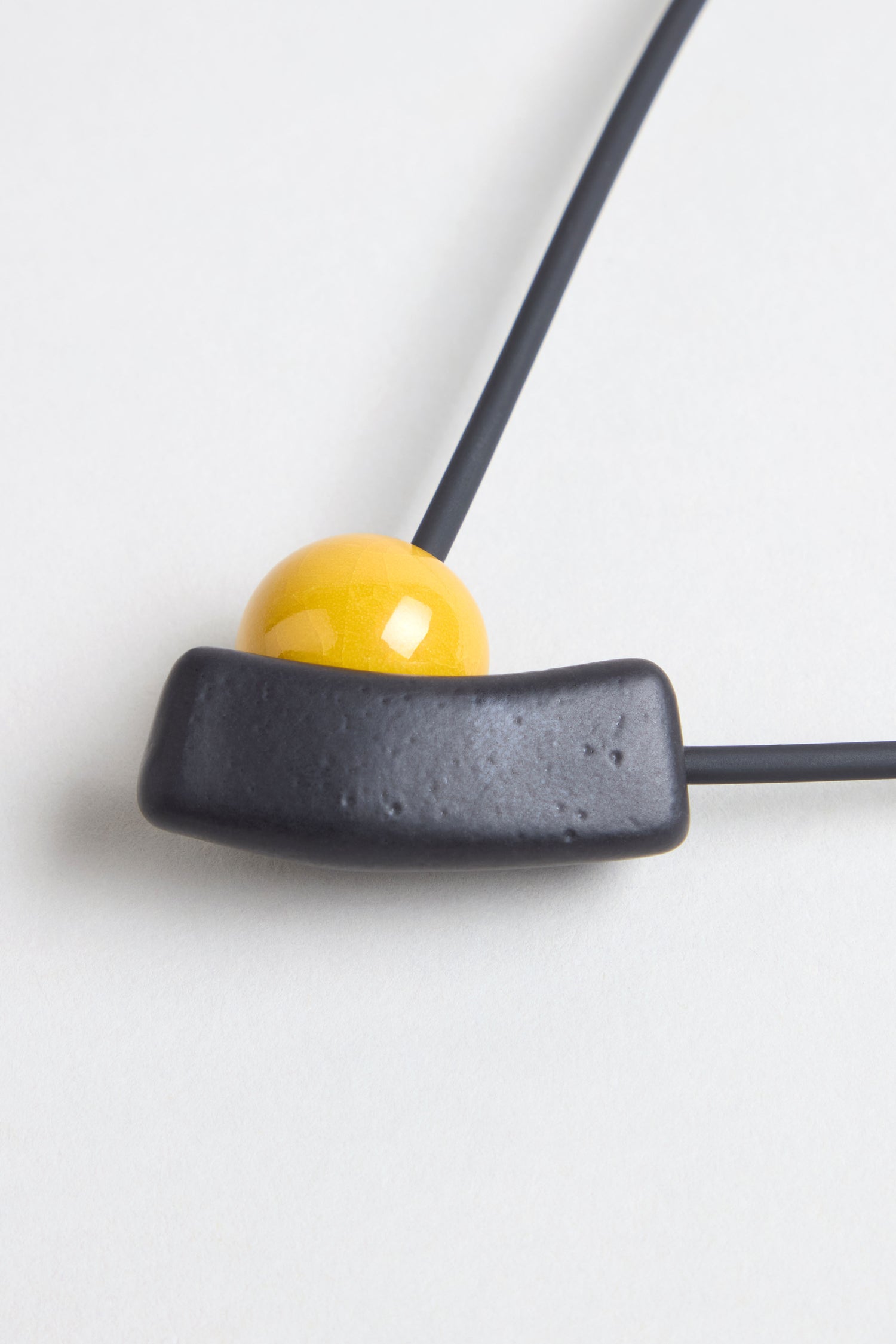 Close-up of the Atom Necklace, a ceramic piece with geometric design, featuring a yellow enamel spherical bead and a black curved rectangular pendant on a black cord, shown against a white background.