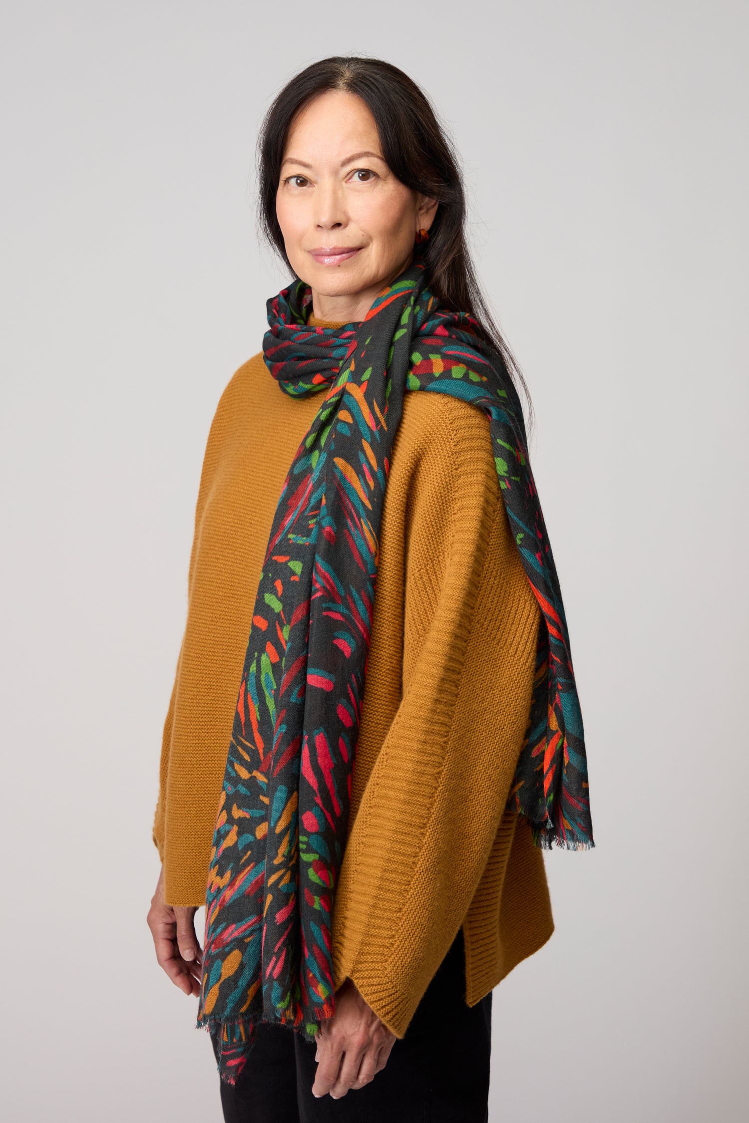A woman with long dark hair wears a mustard yellow sweater and the Skyfire Scarf, standing against a plain light gray background.