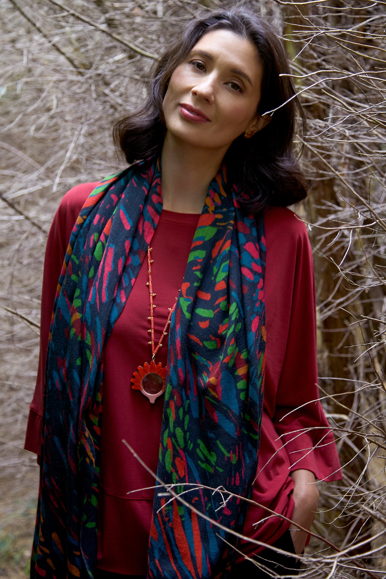 A woman in a red top stands among leafless branches, wearing the Skyfire Scarf and looking at the camera with a neutral expression.