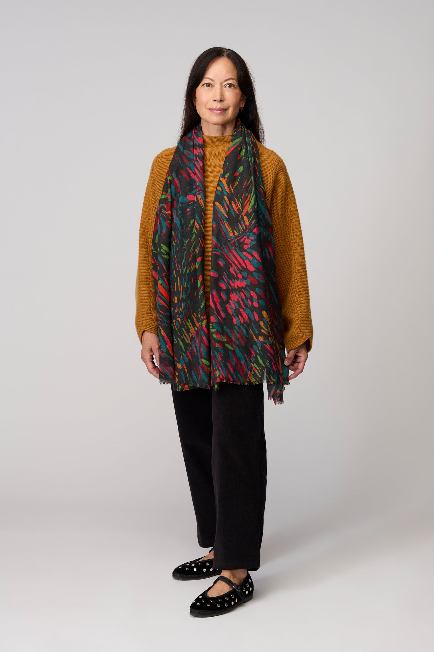 A woman stands against a plain background wearing a mustard sweater, black pants, dotted black shoes, and the Skyfire Scarf, which adds a touch of luxury to her look.