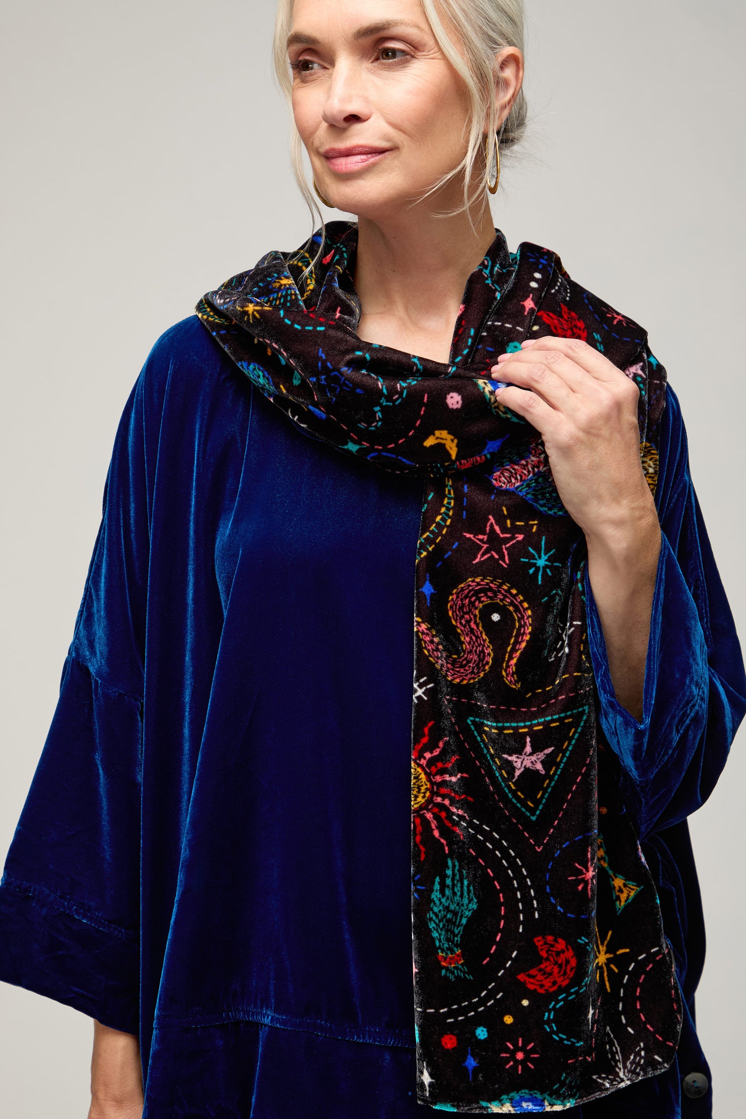 A woman with gray hair wears a royal blue velvet top and the Dream Stitched Print Scarf, featuring artisanal embroidery and colorful patterns, standing against a plain background.