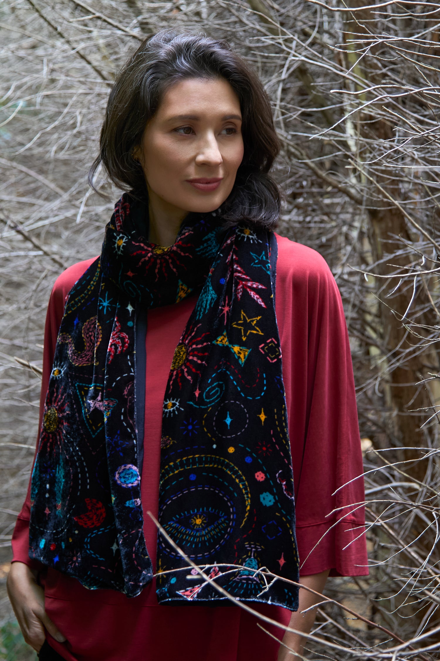 A woman in a red top wears the Dream Stitched Print Scarf—black with vibrant artisanal embroidery—as she stands among leafless branches.