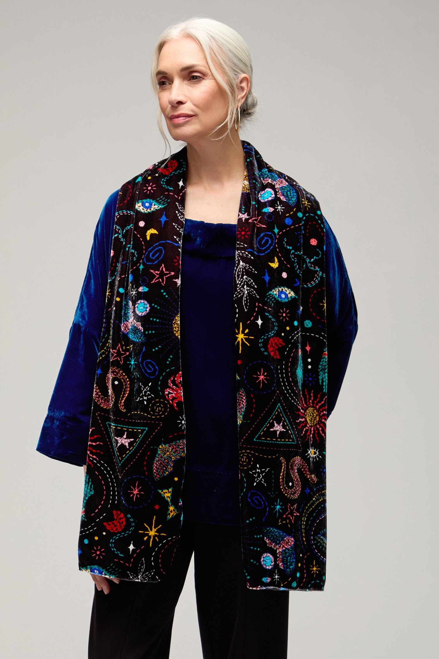 A woman with gray hair in a loose bun wears a blue velvet top and the Dream Stitched Print Scarf, a long, colorful piece featuring artisanal embroidery, against a plain background.
