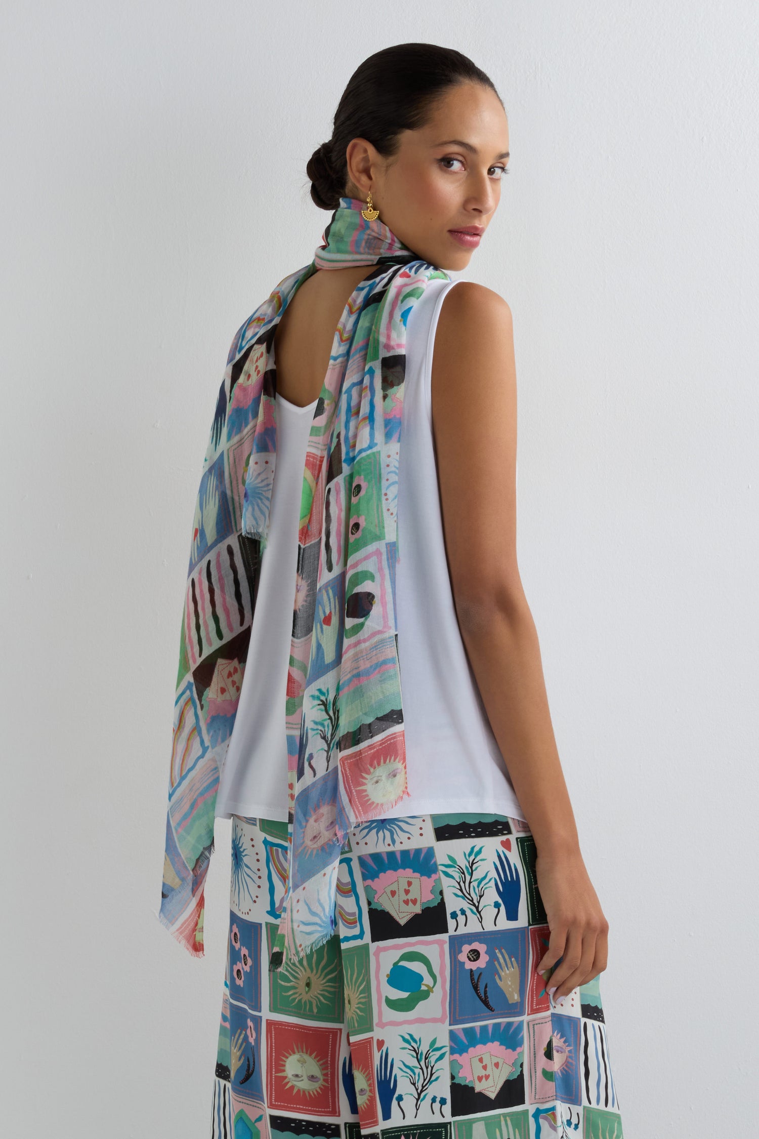 A woman with dark hair in a bun wears a white sleeveless top, matching skirt, and the Mystical Collage Scarf, featuring a storytelling print, as she stands against a plain white wall.