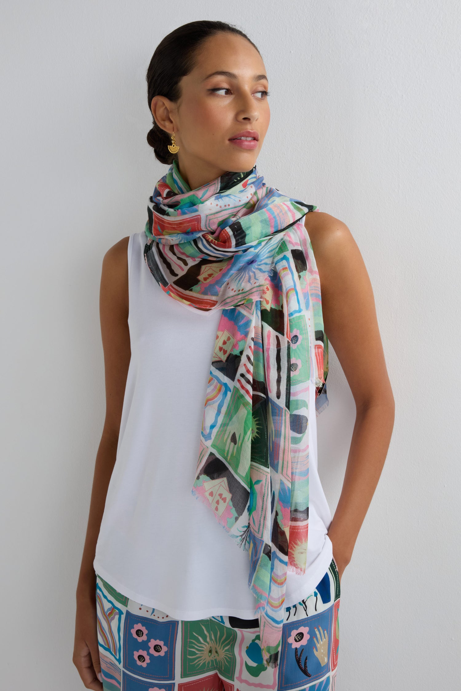 A woman stands against a white wall, looking to the side, styled in a white sleeveless top, patterned pants, and the Mystical Collage Scarf featuring a storytelling print.