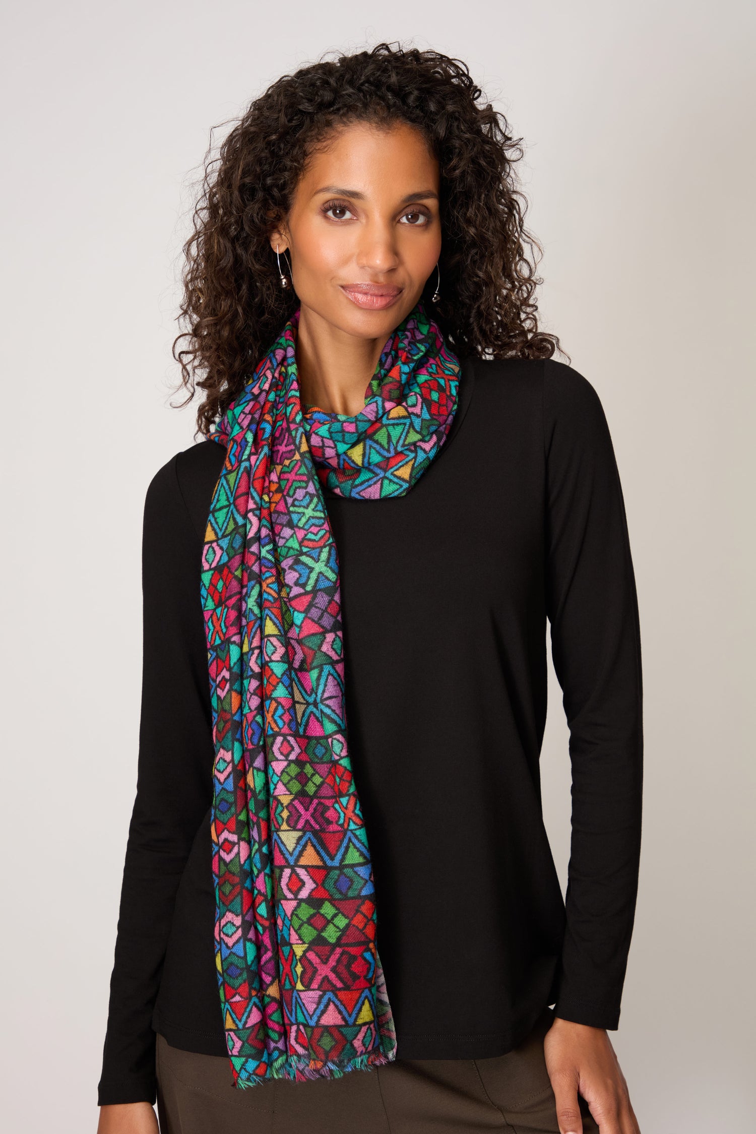A woman in a black long-sleeve top wears the Peruvian Stitched Print Scarf, featuring traditional Peruvian patterns and vibrant colors, against a light background—a perfect lightweight accessory.