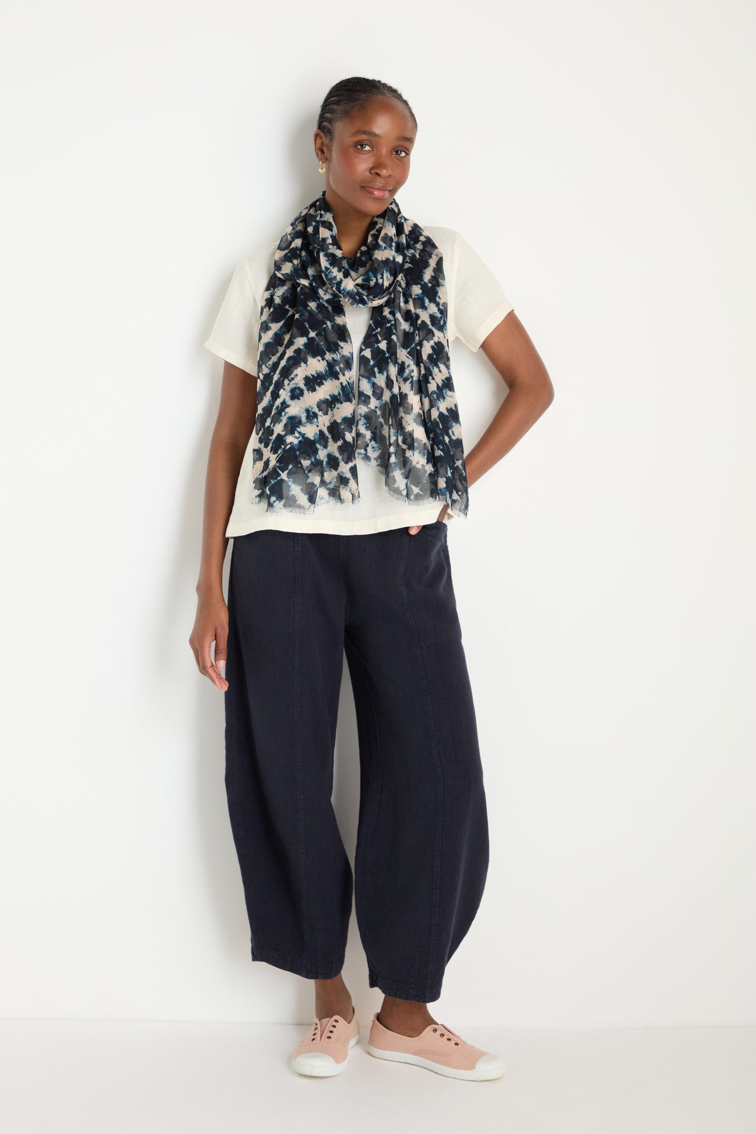 A woman stands against a white wall in a white t-shirt, dark loose pants, and light pink sneakers, her look finished with the Diamond Tie Dye Scarf for a standout touch.