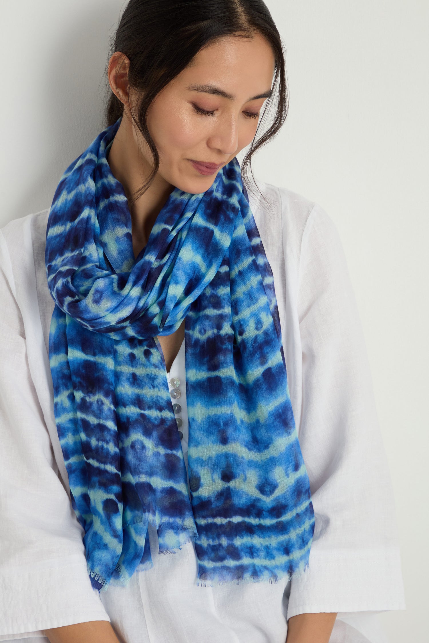 A woman in a white top wears the Tie Dye Print Scarf in blue modal silk, looking down with a neutral expression against a plain background.
