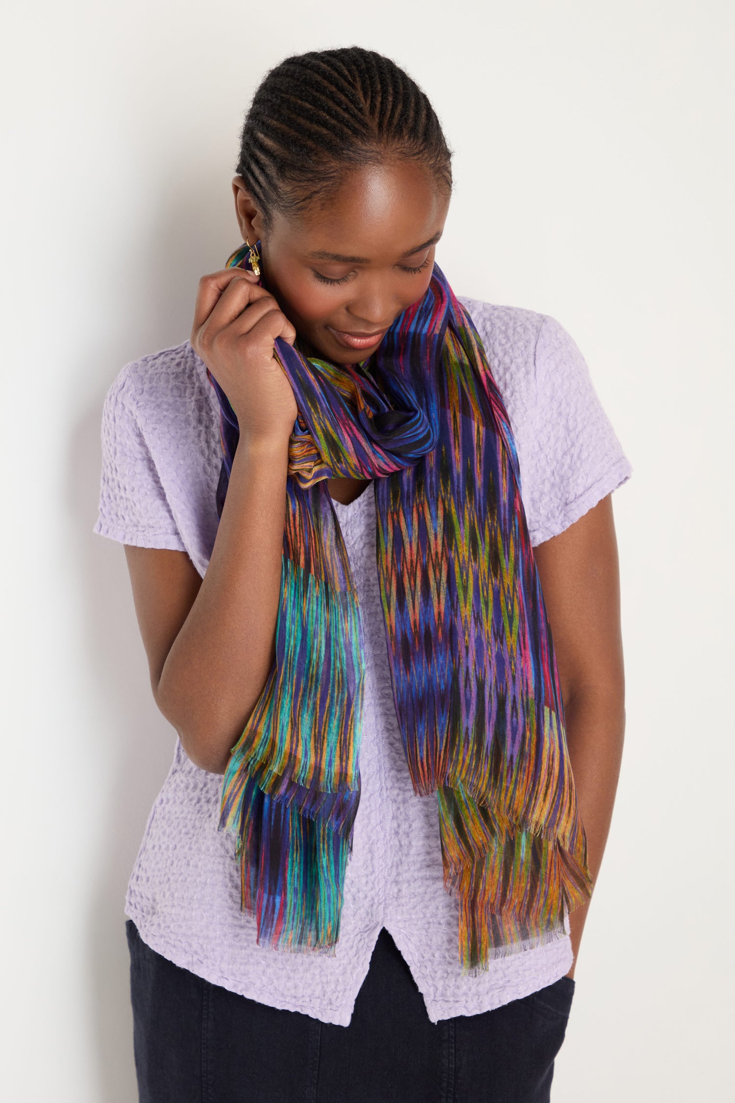 A woman in a light purple textured blouse looks down and touches her Vivid Ikat Patchwork Scarf, showcasing its vibrant multicolored design.