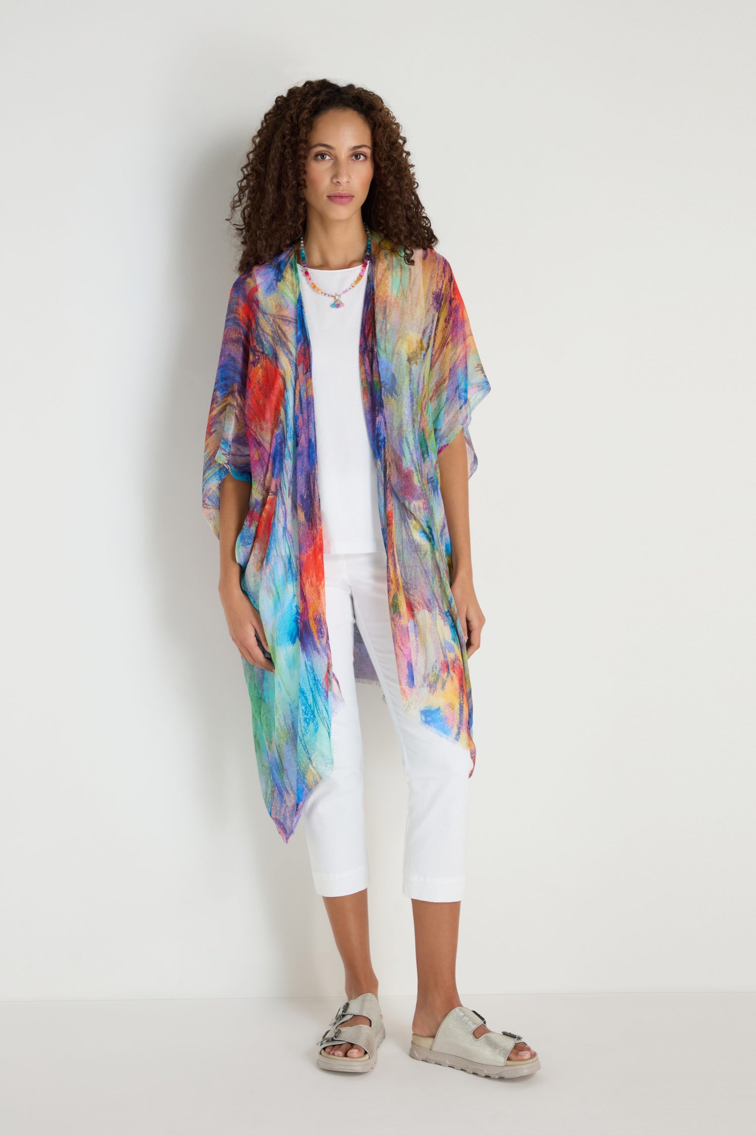 A woman stands against a plain wall wearing a white outfit, the Soft Meadow Dreams Kimono draped over her shoulders, beige sandals, and a necklace.
