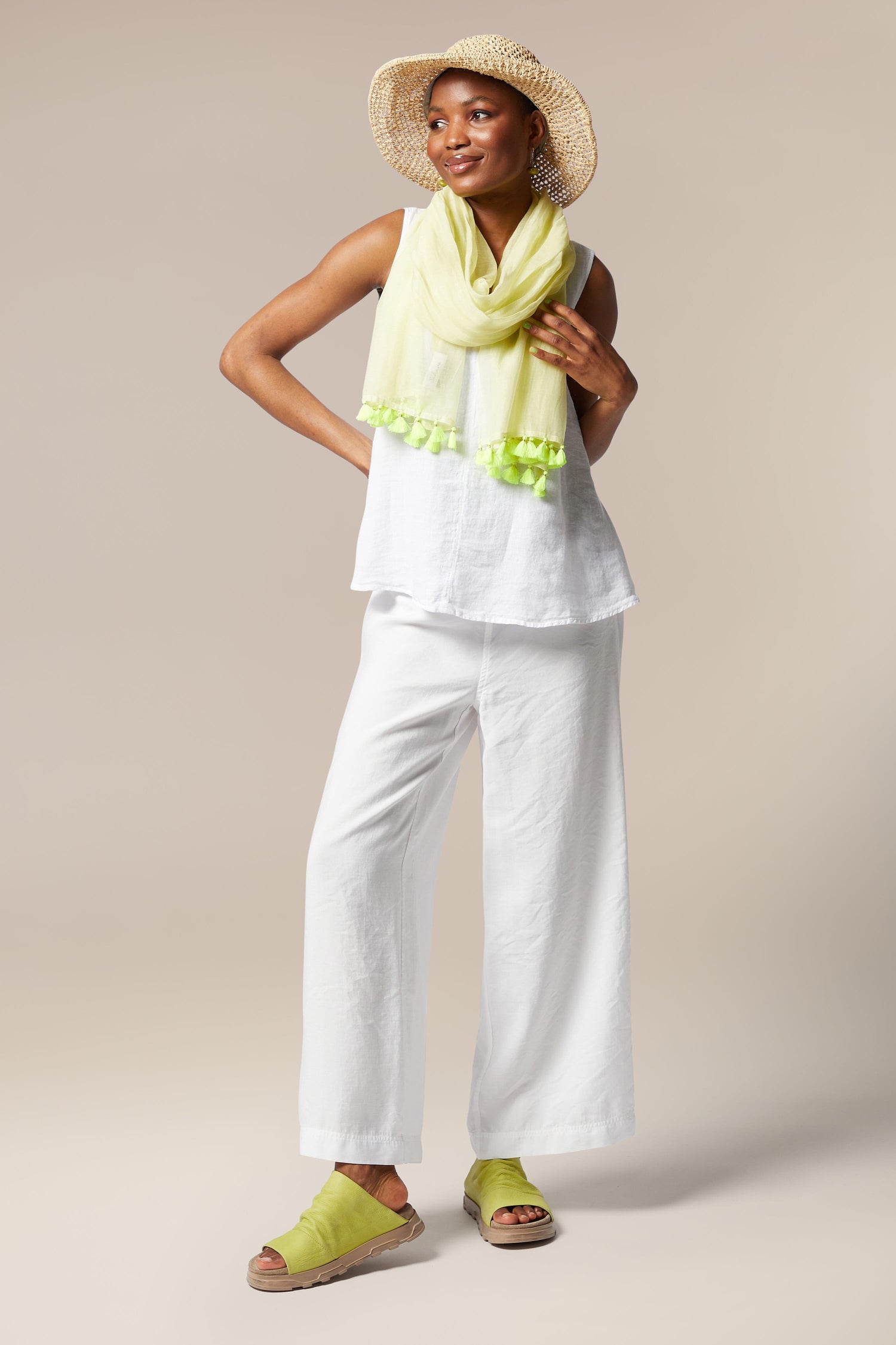 A woman in a summer outfit stands confidently, wearing a white sleeveless top, white pants, Neon Tassel Scarf, sandals, and a straw hat against a beige background.