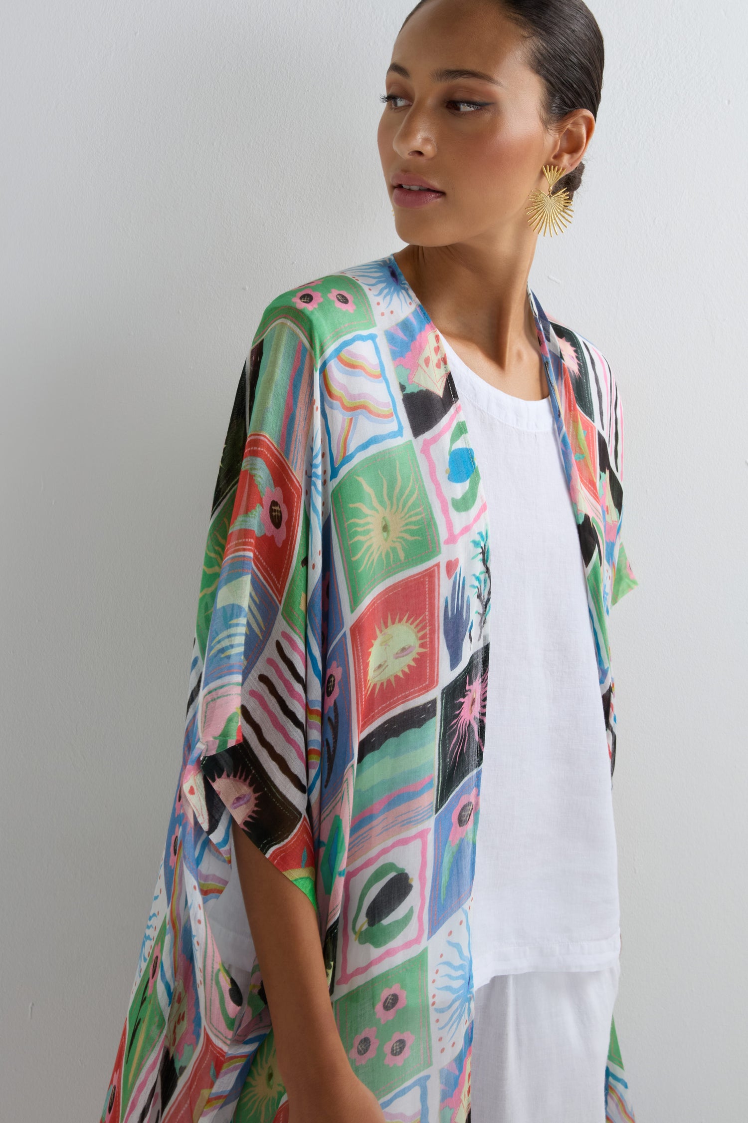 A woman in the Mystical Collage Throwover, a vibrant patterned kimono, stands against a light background in a white outfit—ideal for making a statement at summer events.