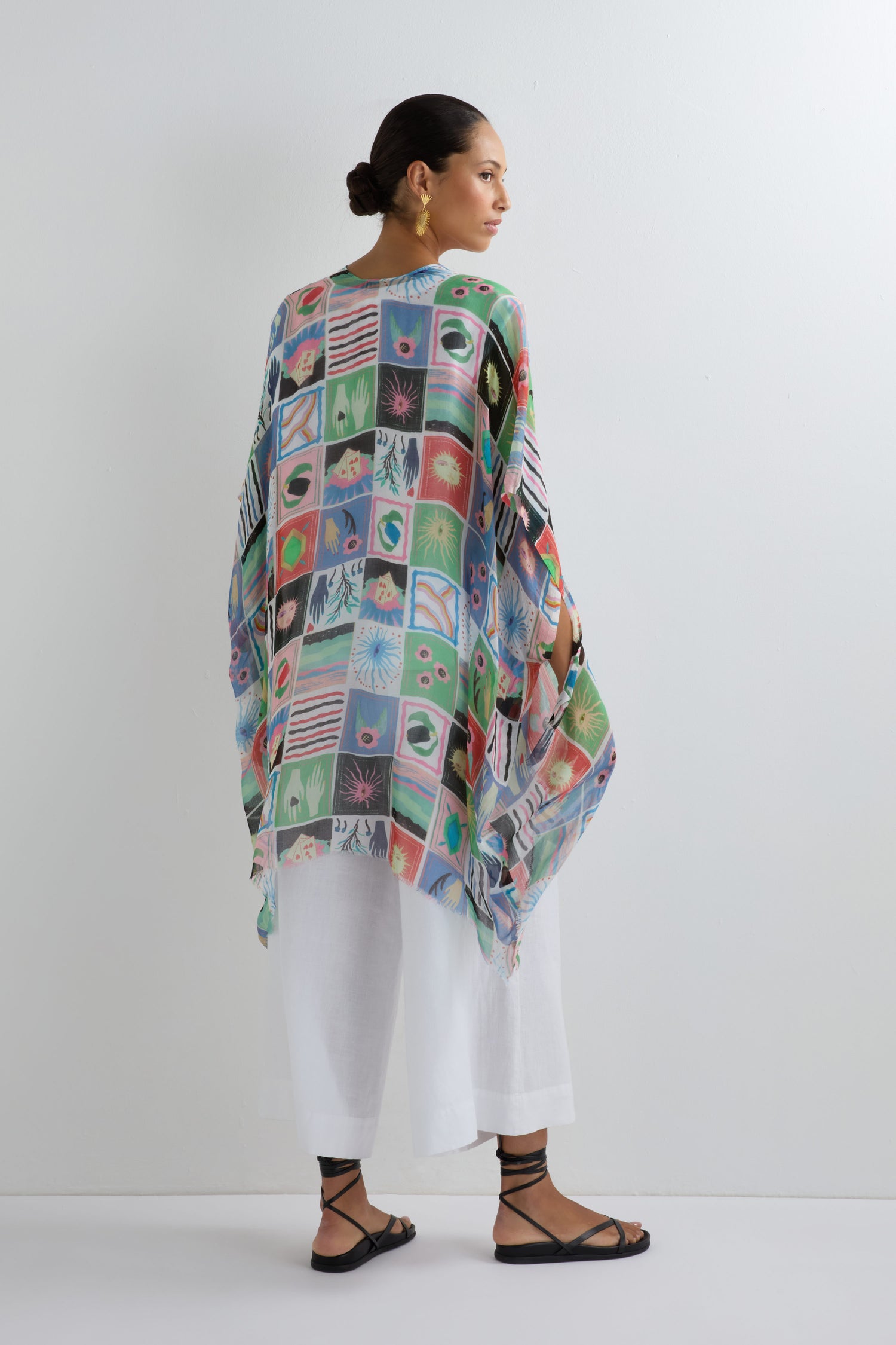 A person faces away in the Mystical Collage Throwover, a vibrant patterned cape that adds a statement to summer looks, styled here with a white outfit and black sandals against a plain white background.