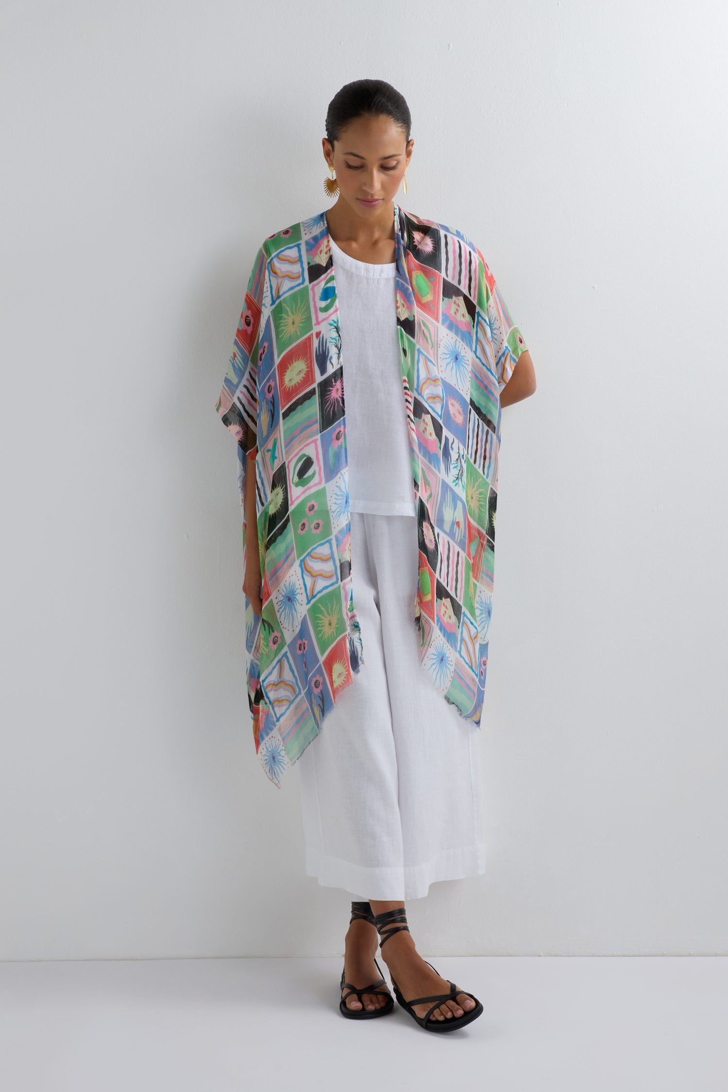 A woman poses by a white wall in a white outfit, layered with the Mystical Collage Throwover—a vibrant, patterned kimono—making a bold statement for summer events, paired stylishly with black sandals.