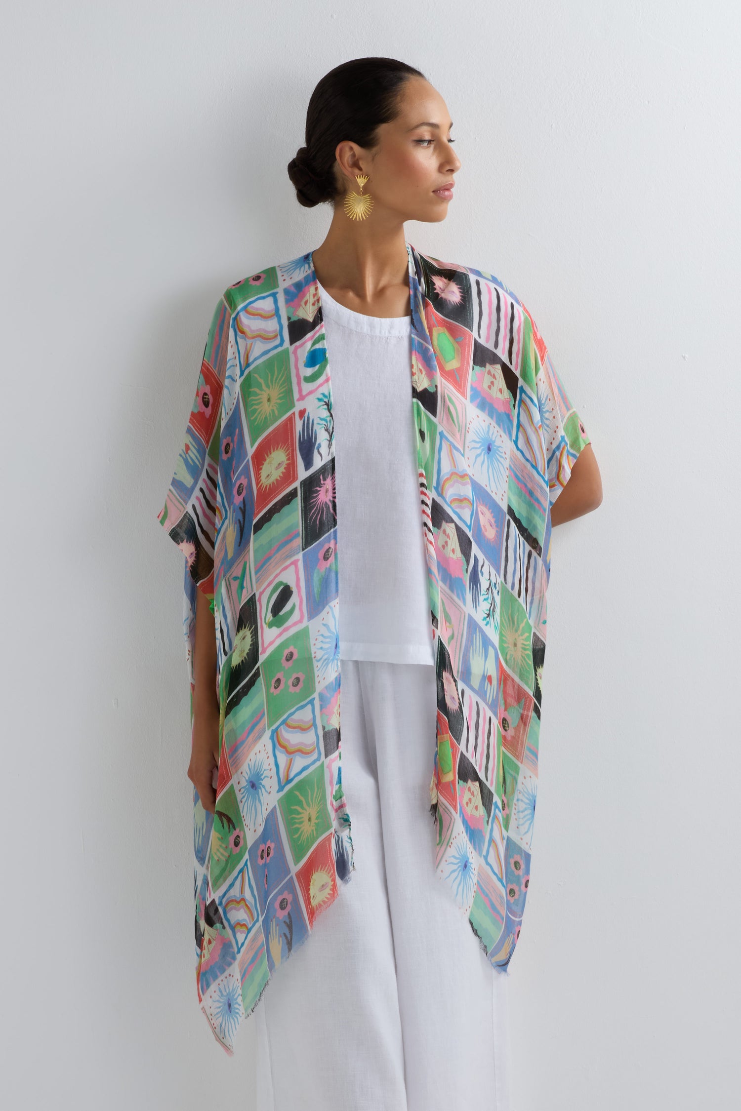 A woman wearing a white outfit and the Mystical Collage Throwover—a vibrant, patterned open-front kimono—stands against a plain white wall, embodying bold style as she looks to the side.