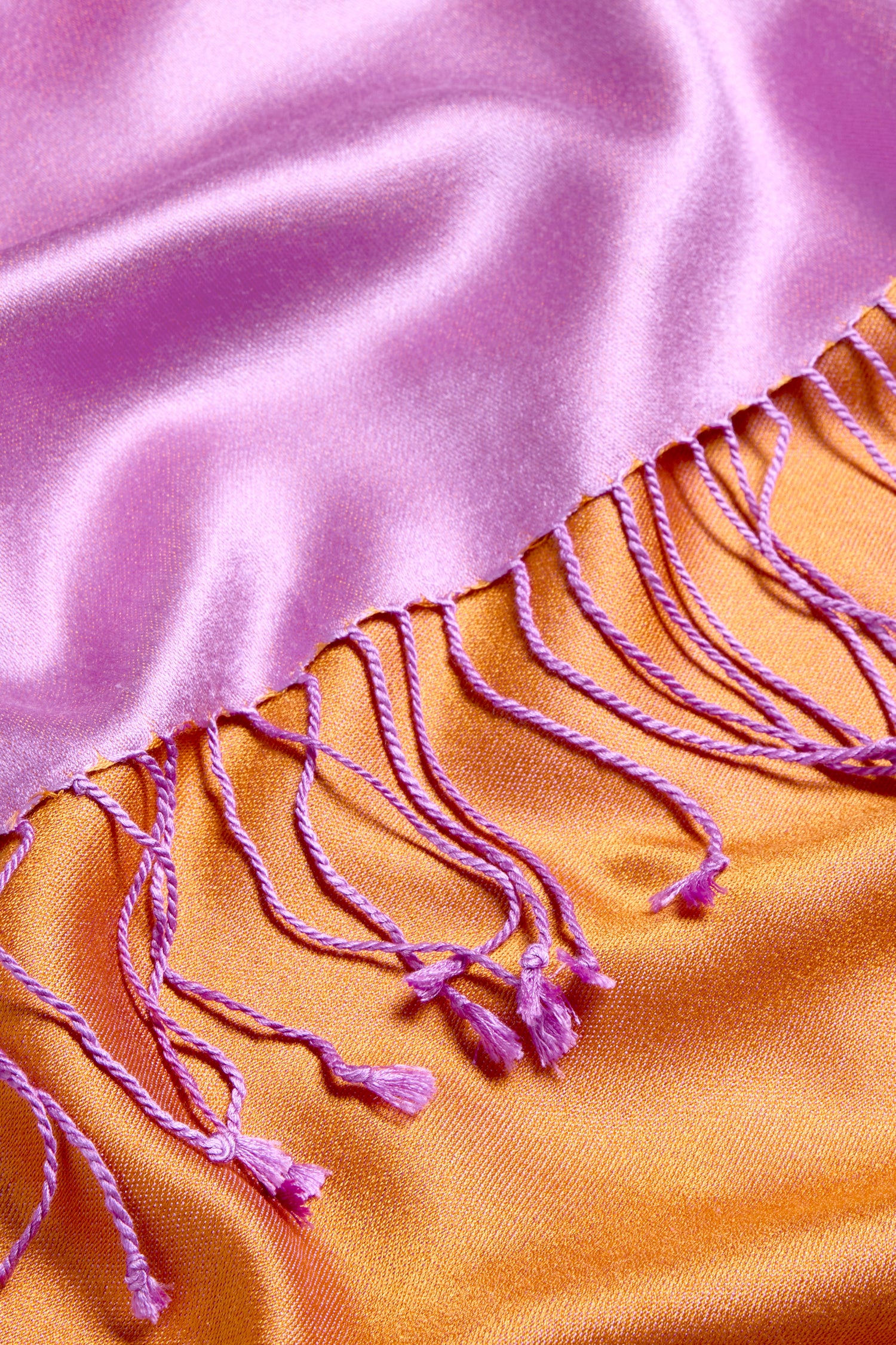 A close-up of the Two Tone Silk Scarf showcases its pink and orange colourblocked design, highlighted by pink twisted fringe along the edge and a luxurious sheen.