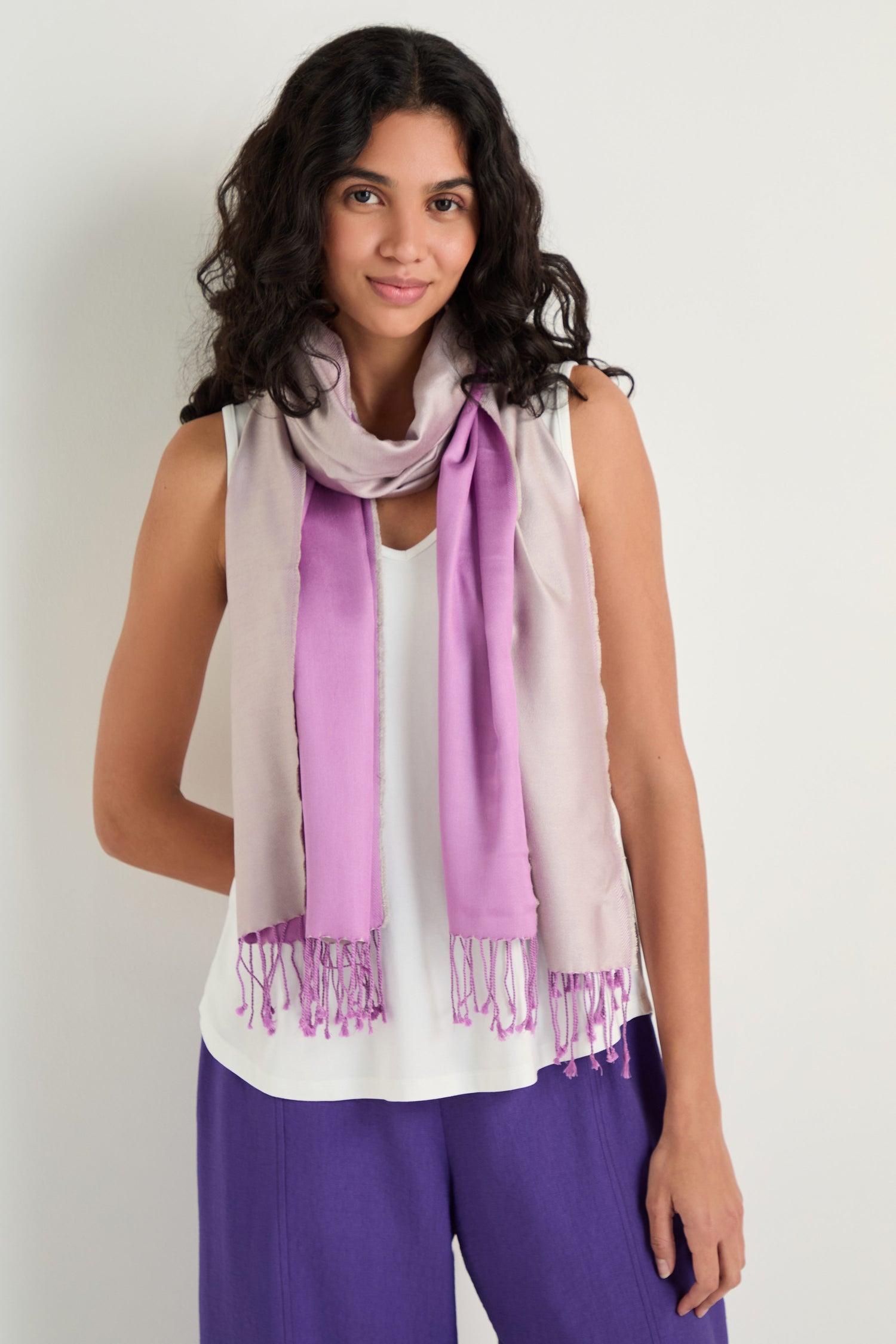 A woman with long curly hair wears the Two Tone Silk Scarf in light purple, draped over a white sleeveless top and purple pants, standing against a plain light background.