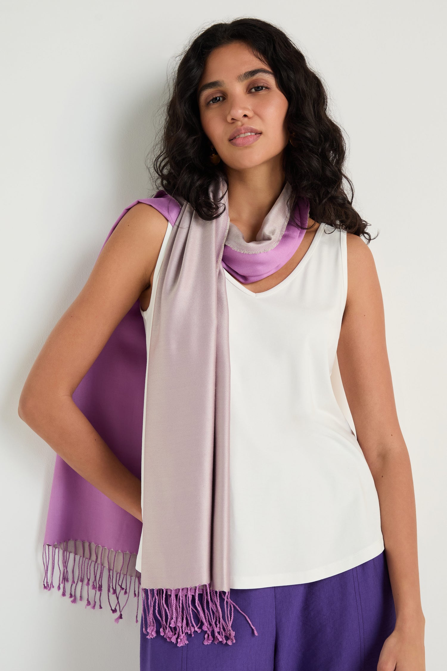 A woman with long curly hair wears a sleeveless white top, purple pants, and the Two Tone Silk Scarf in lavender and purple, standing against a plain light-colored wall.