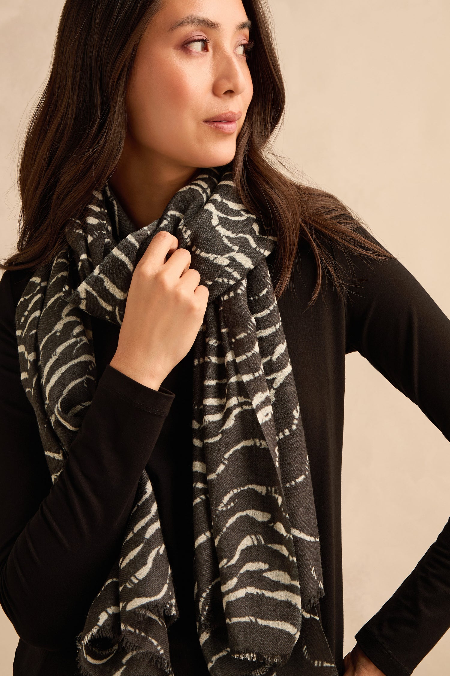 A woman wearing a black long-sleeve top drapes the Dancing Waves Scarf, featuring a striking black-and-white wave pattern, and gazes to the side while holding the scarf.