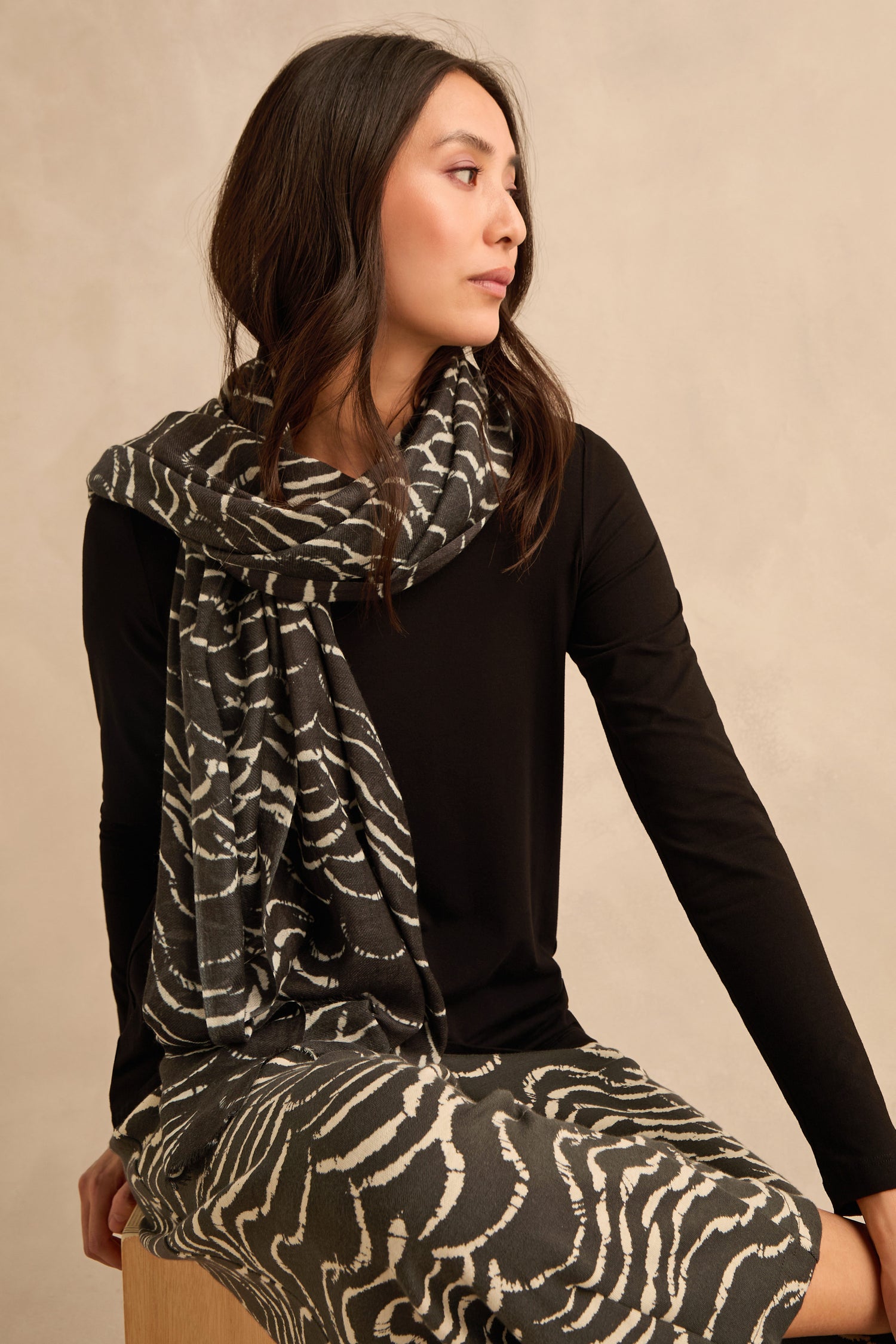 A woman sits on a wooden stool, wearing a black long-sleeve shirt and the Dancing Waves Scarf—a lightweight winter accessory featuring a striking wave pattern—paired with a matching skirt, looking to the side against a beige background.