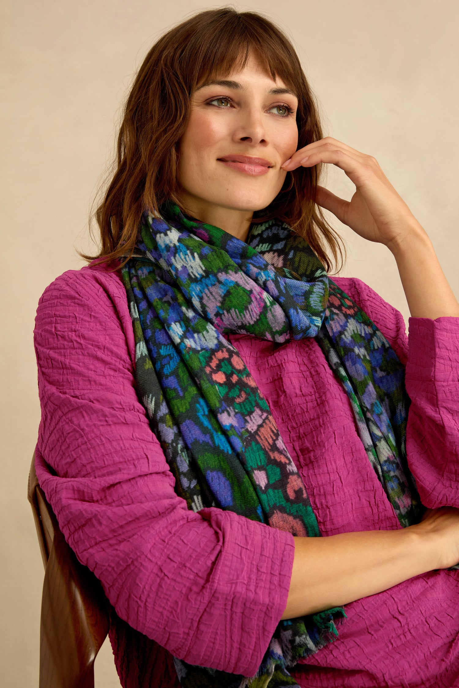 A woman in a magenta textured top and the Winter Garden Wool Scarf sits on a wooden chair, resting her hand on her face and smiling slightly.