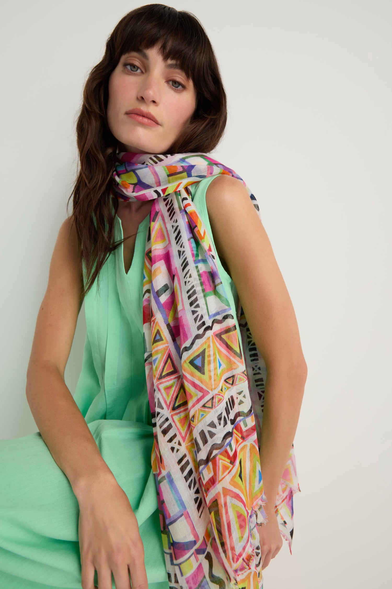 A woman in a sleeveless mint green dress sits against a plain light background, draping the Painted Geometric Scarf—featuring colorful geometric patterns—over her shoulder.
