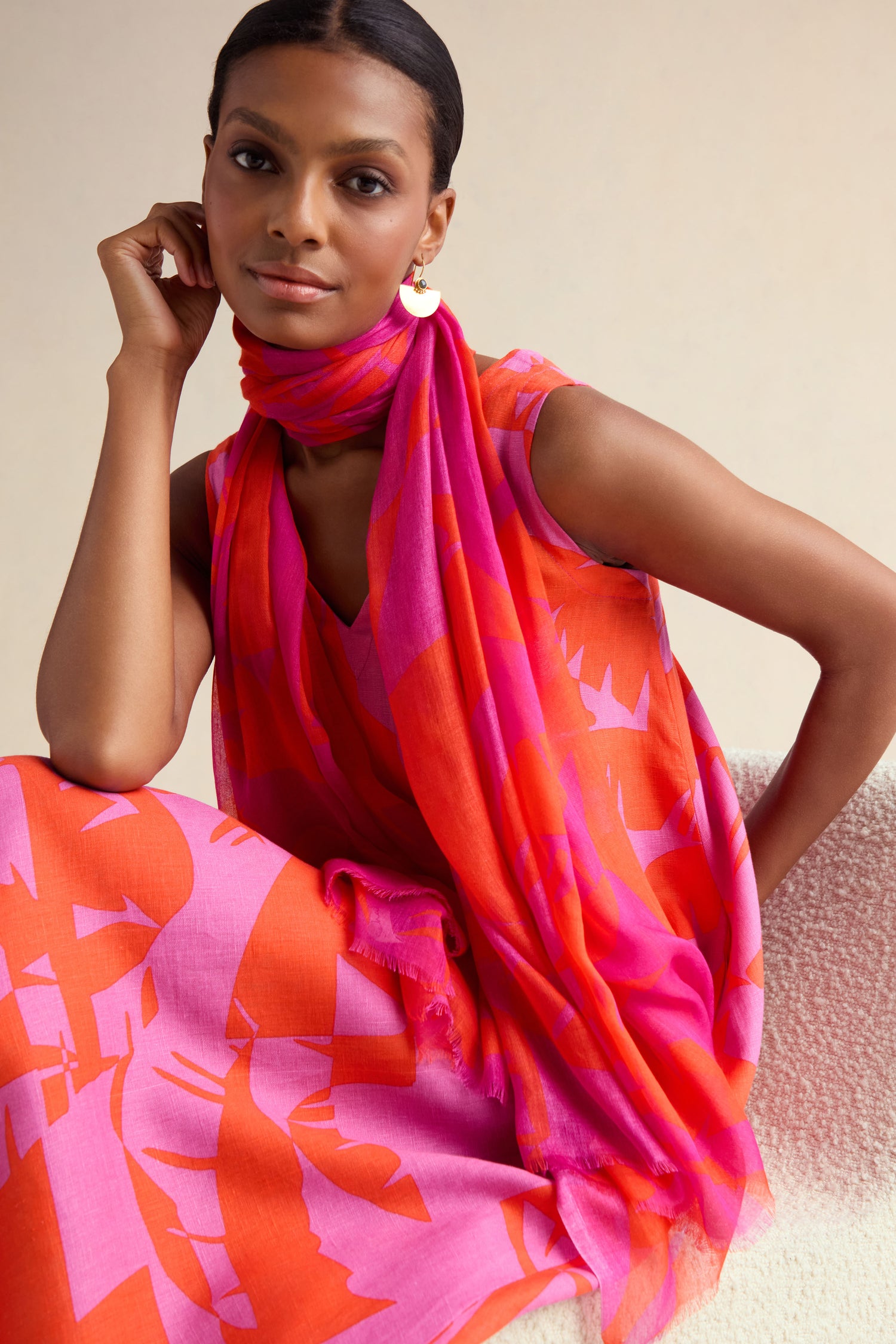 A woman wearing a sleeveless pink and orange patterned dress with the Palm Mirage Scarf sits on a textured chair, looking at the camera.