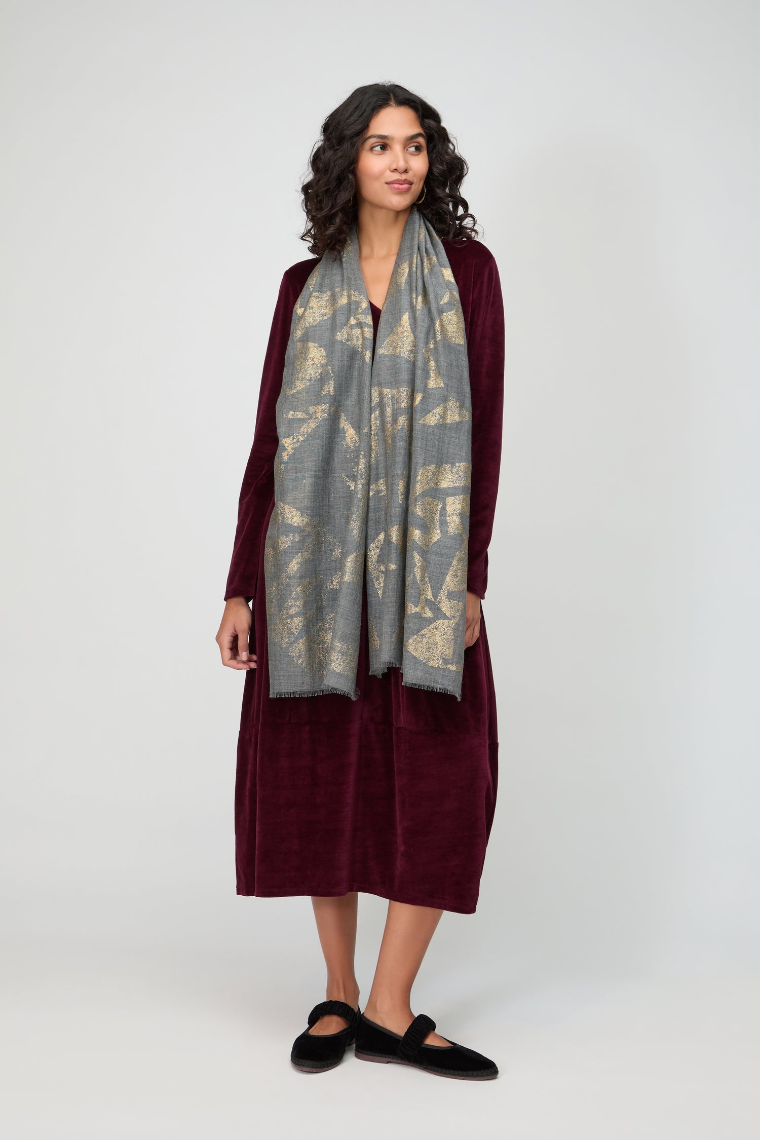 Woman stands against a plain background, wearing a long burgundy dress, the Aurelian Cotton Scarf in gray, and black slippers.