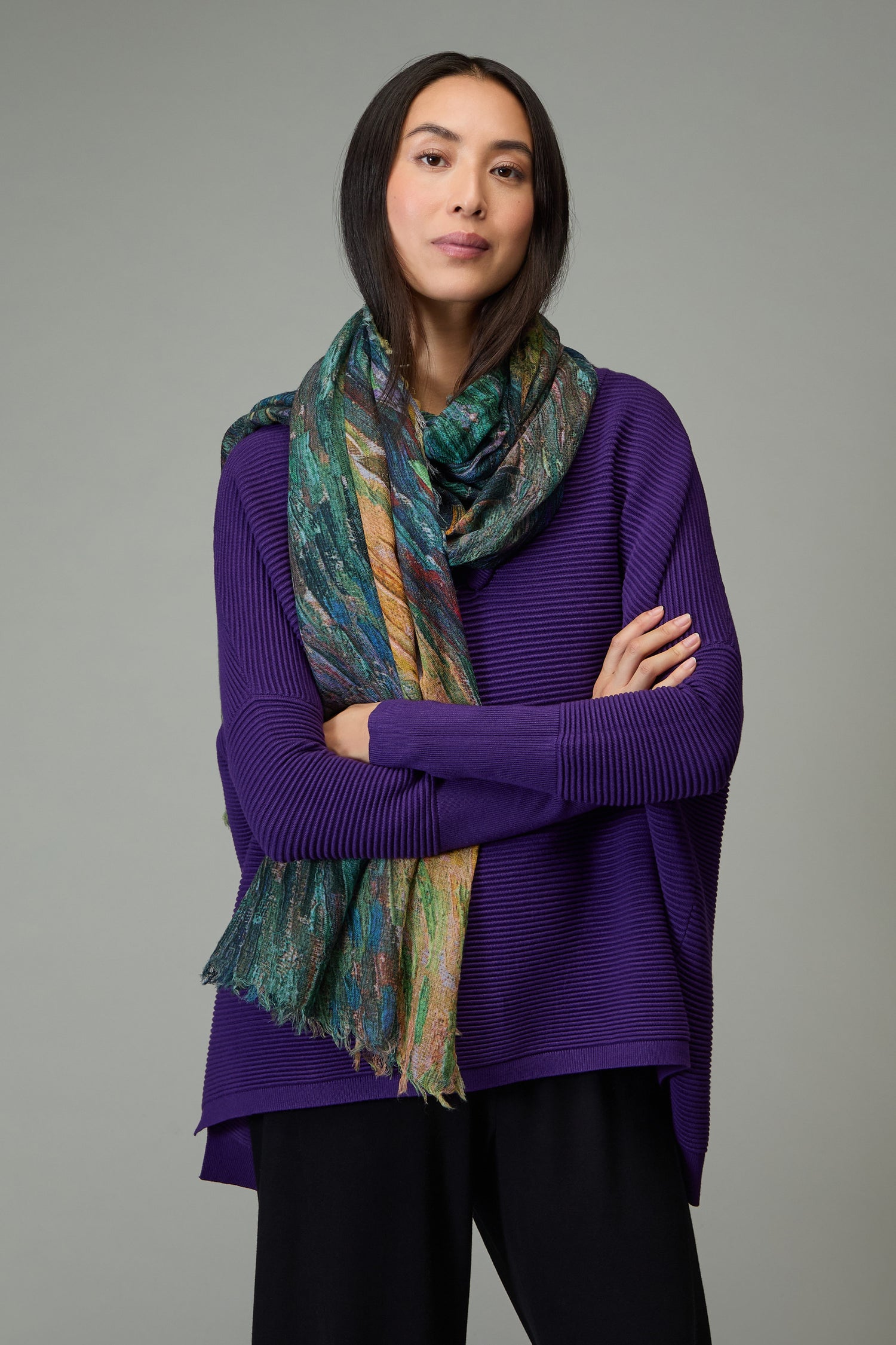 A woman with arms crossed wears a textured purple sweater, black pants, and the Painter's Landscape Wool Scarf—an abstract print accessory—against a plain gray background.