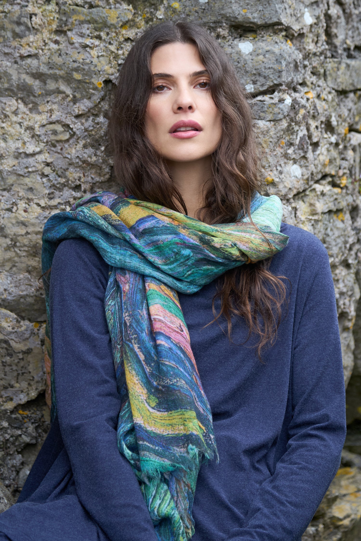 A woman with long brown hair, wearing a blue top and the Painter's Landscape Wool Scarf, sits in front of a stone wall.
