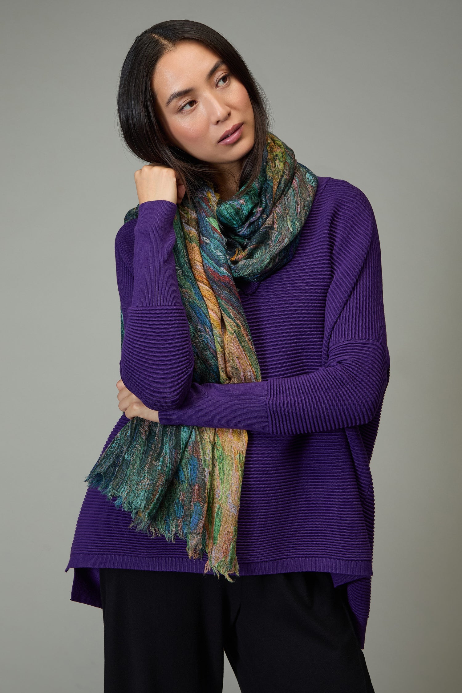 A woman in a purple sweater models the Painter's Landscape Wool Scarf, featuring an abstract print, as she poses with one hand near her face against a plain gray background.