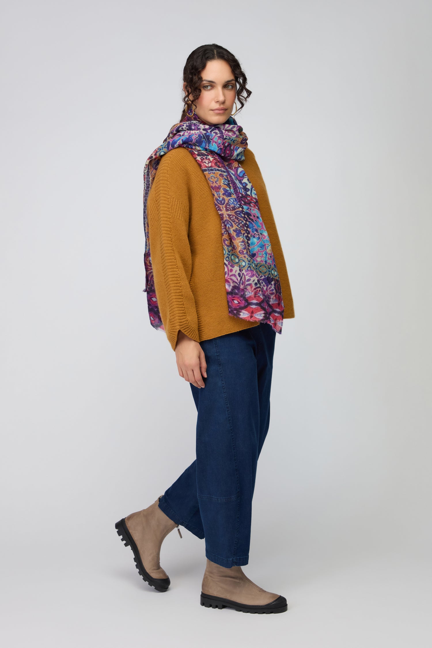 A person stands in profile against a plain light background, wearing a mustard sweater, blue jeans, tan boots, and the Patchwork Mural Print Wool Scarf.