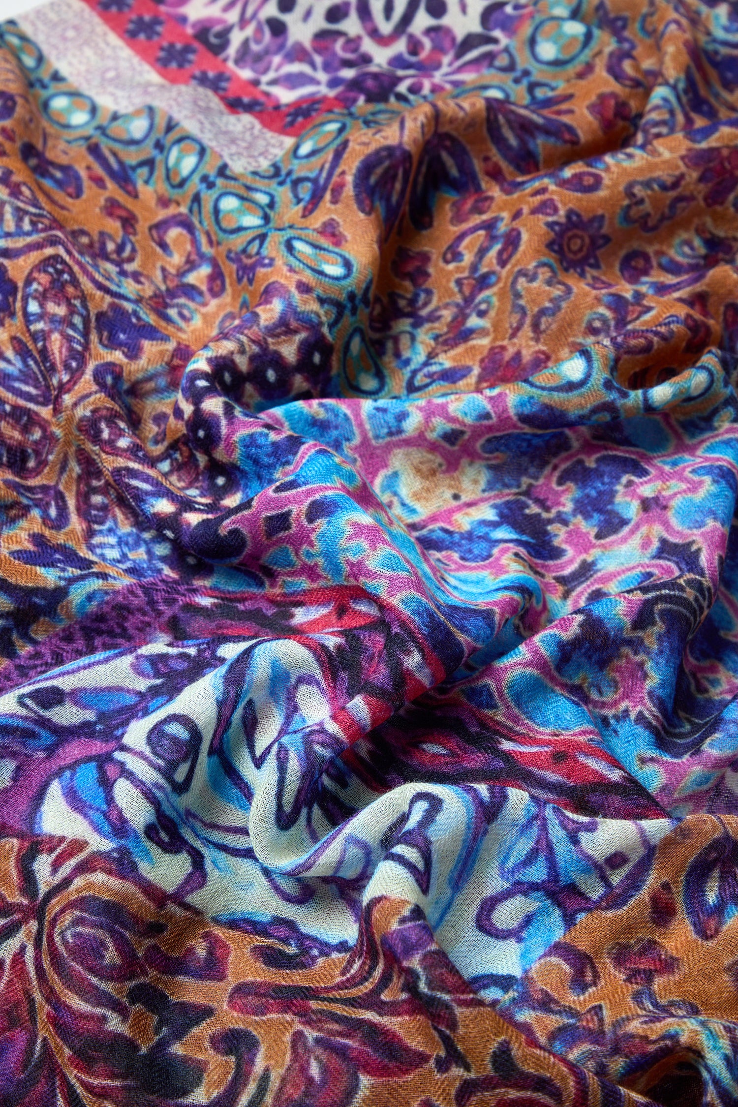 The Patchwork Mural Print Wool Scarf features a vibrant mix of blue, purple, orange, and pink in intricate patterns, shown draped in soft folds for a colorful statement look.