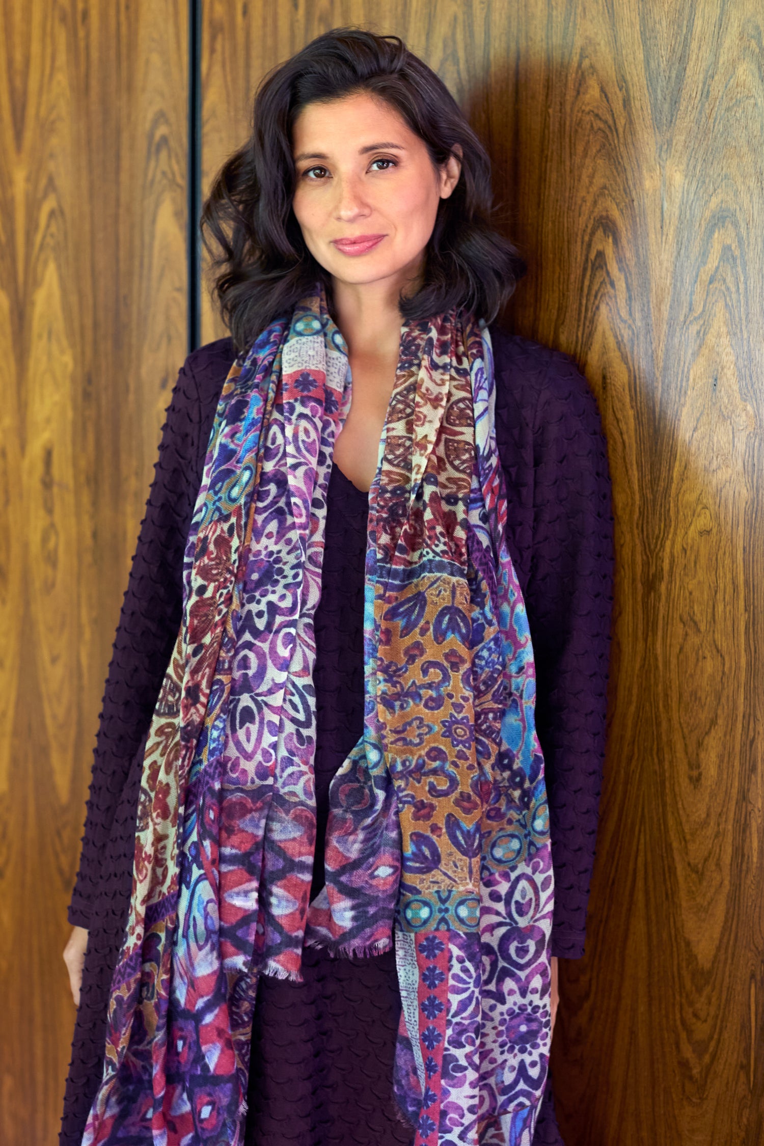 A woman with shoulder-length dark hair stands before a wooden wall, wearing a textured dark purple dress and the Patchwork Mural Print Wool Scarf.