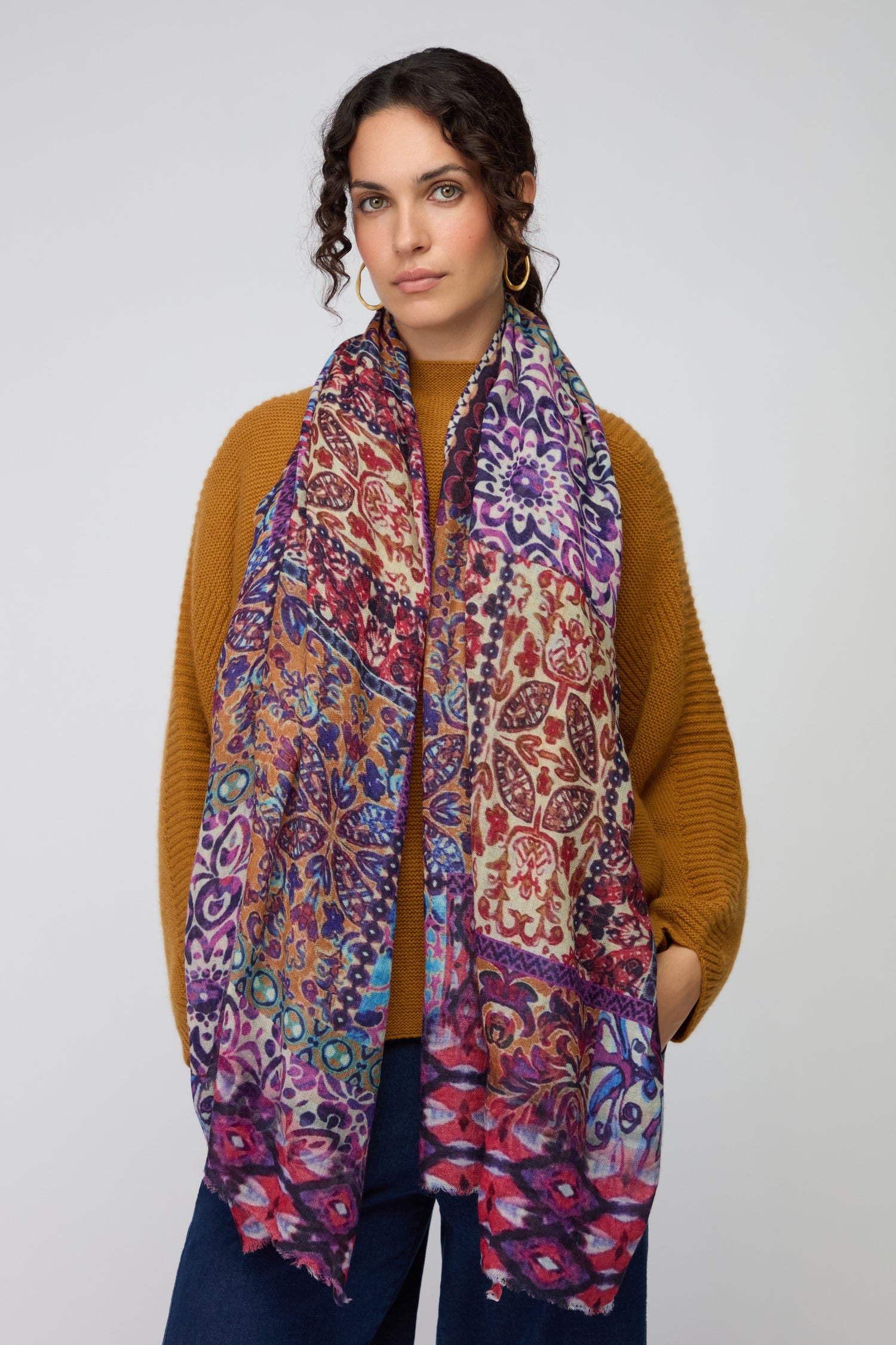 A woman stands against a plain light background with her hands in her pockets, wearing a brown sweater and the Patchwork Mural Print Wool Scarf.
