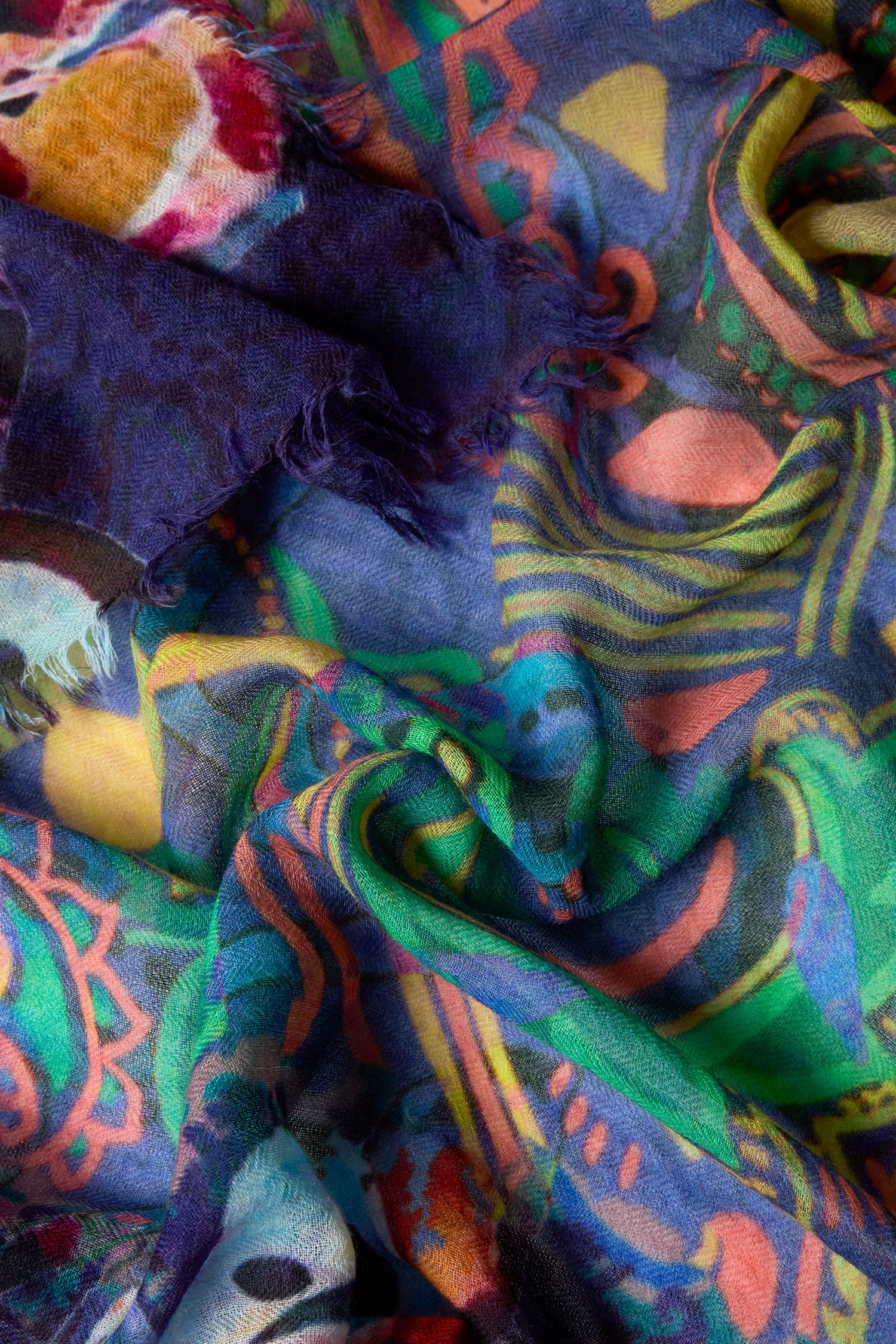 A close-up of the Painted Paisley Wool Scarf shows colorful abstract patterns and frayed edges, crafted from a lightweight wool blend with subtle paisley accents.