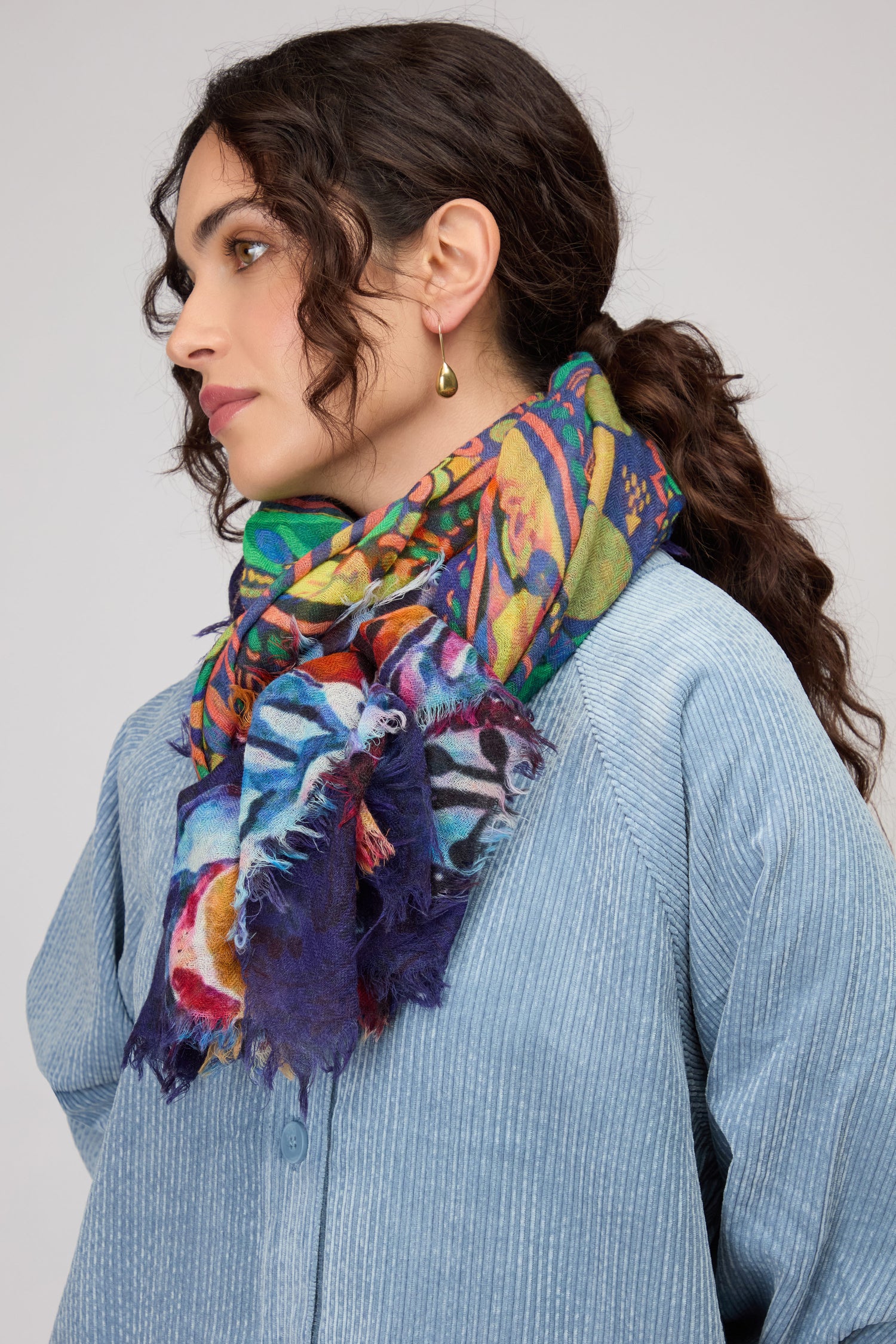 Woman in a blue textured shirt models the Painted Paisley Wool Scarf, featuring a colorful paisley design, and gold earrings, facing left against a plain background.