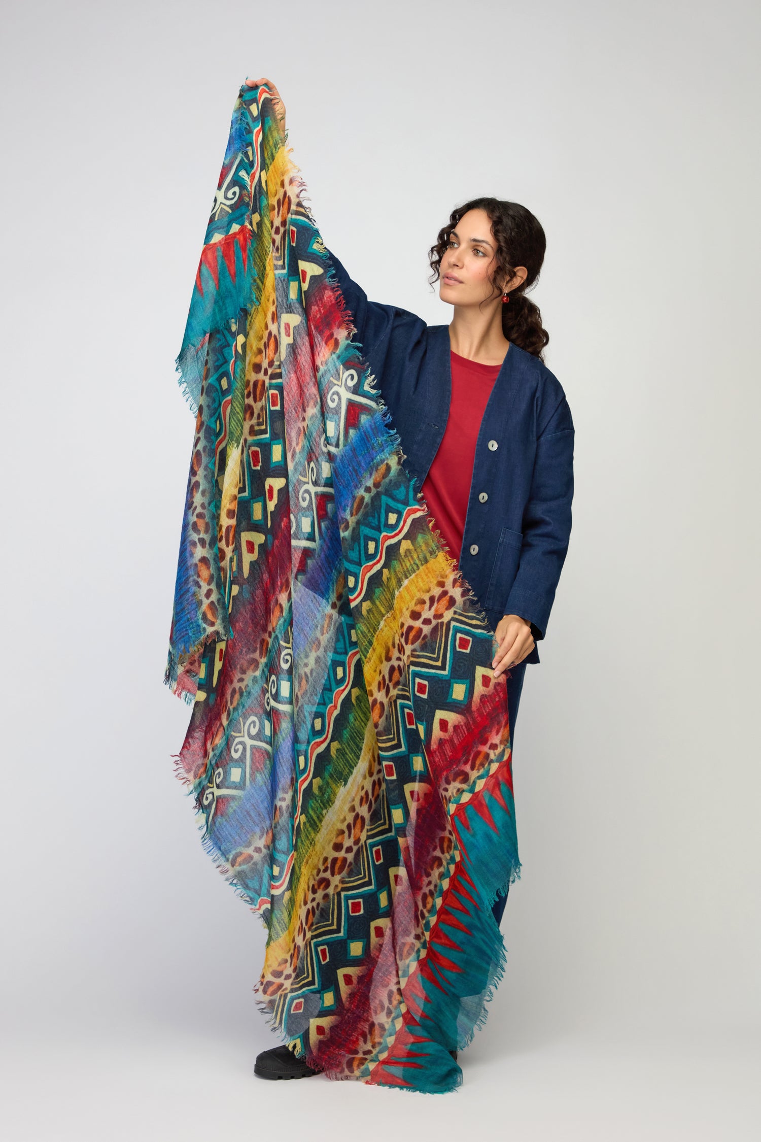 A woman in a blue jacket and red dress displays the Savannah Print Wool Scarf—an oversized, colorful wool wrap with fringed edges—against a plain light background.